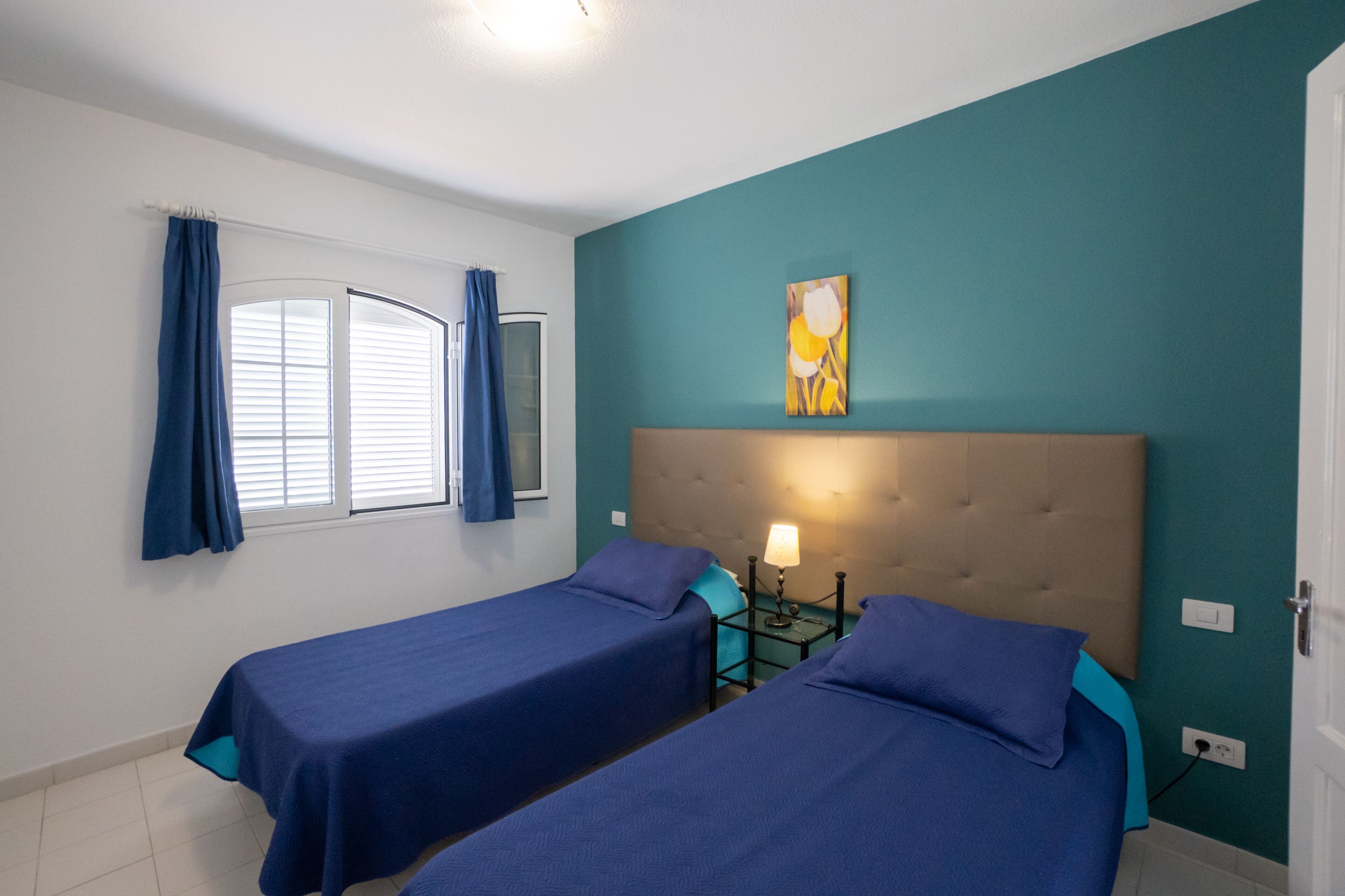 Ein Zimmer mit zwei Betten, einem Vorhangfenster, einem Bild an der Wand und einer Nachttischlampe.