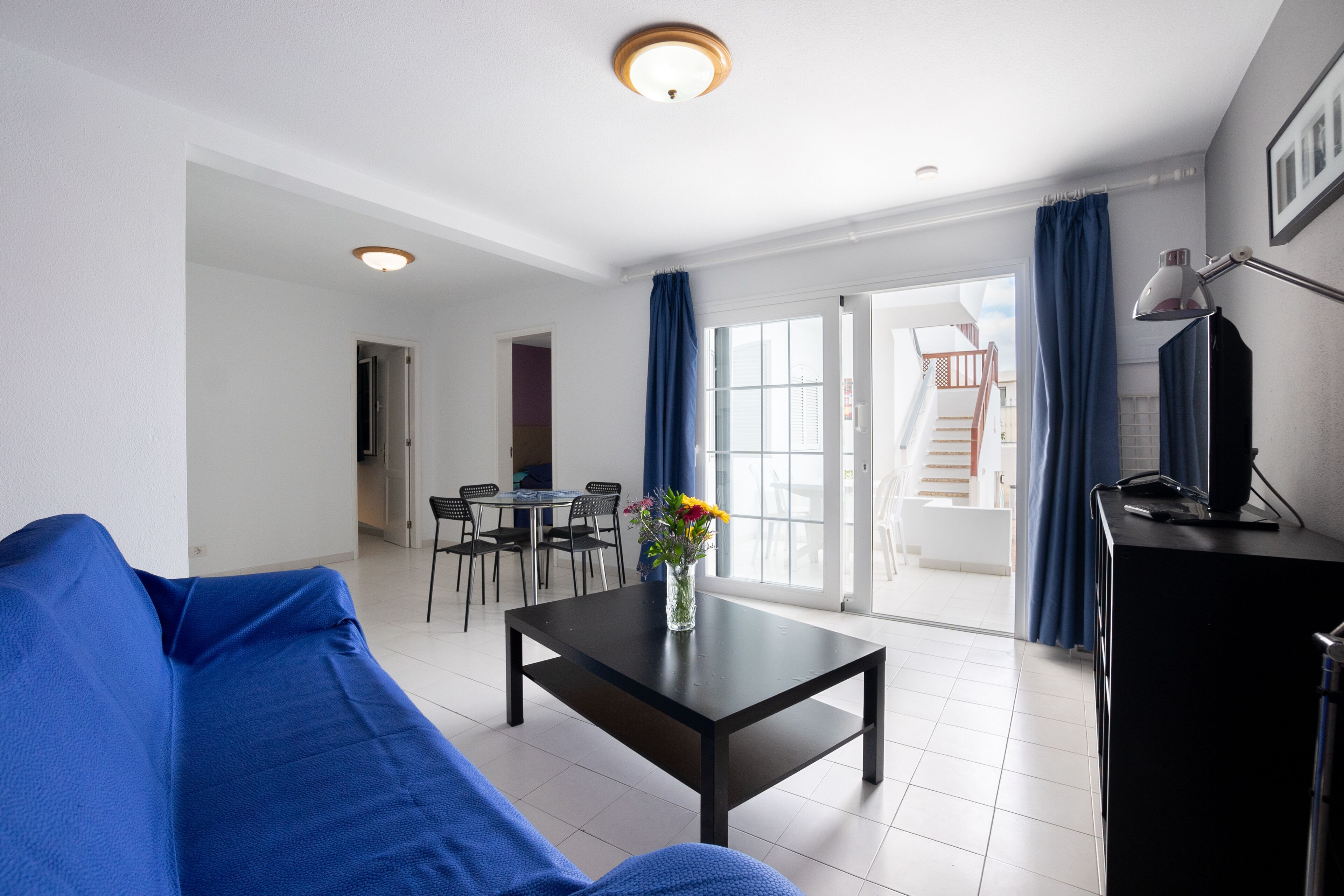 A modern living room with a blue sofa, a coffee table with a vase of flowers, a dining area with chairs, and a staircase leading to an outdoor balcony.