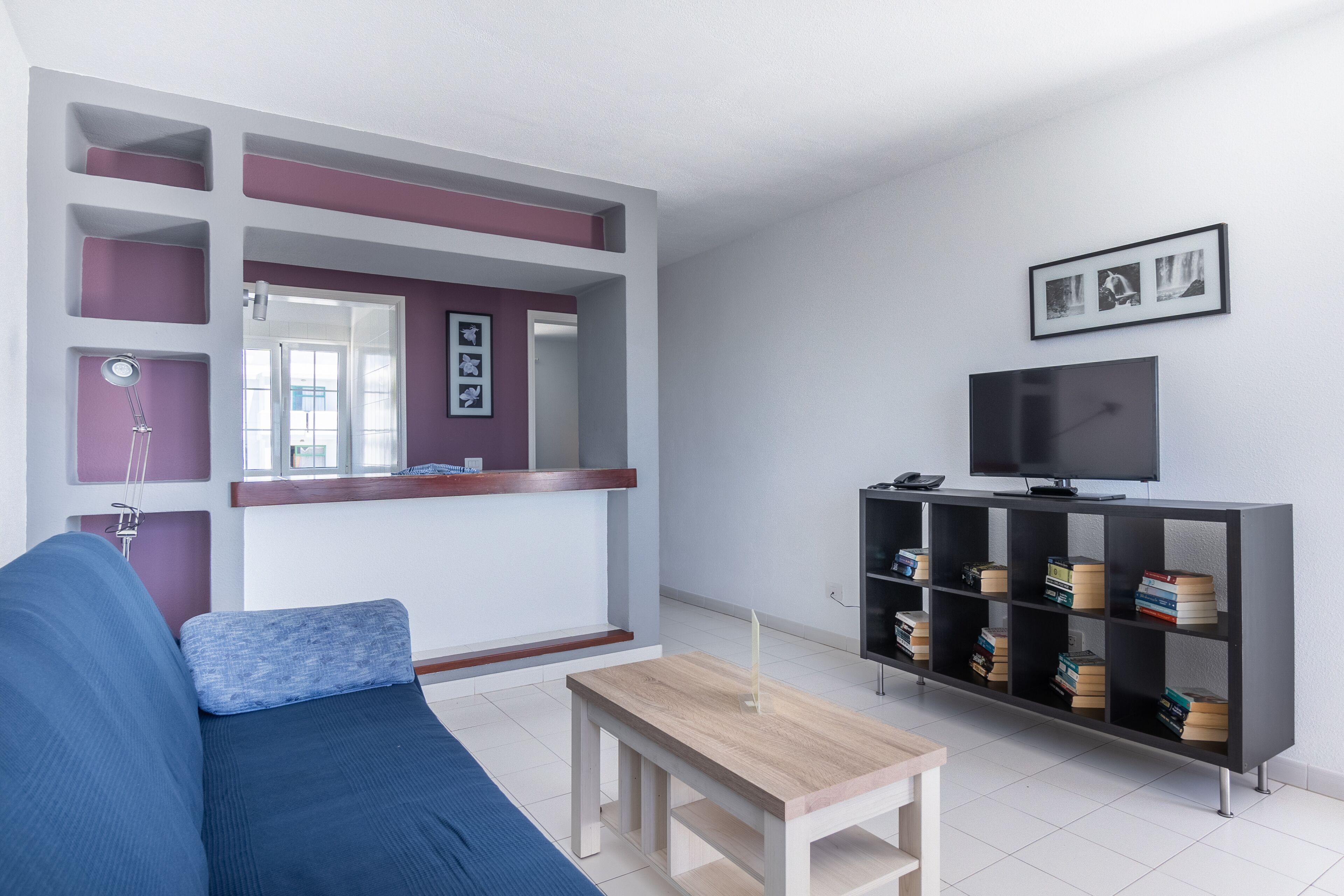 Una sala de estar con un sofá azul, una mesa de centro de madera, un mueble de televisión negro con libros y un espejo en la pared.