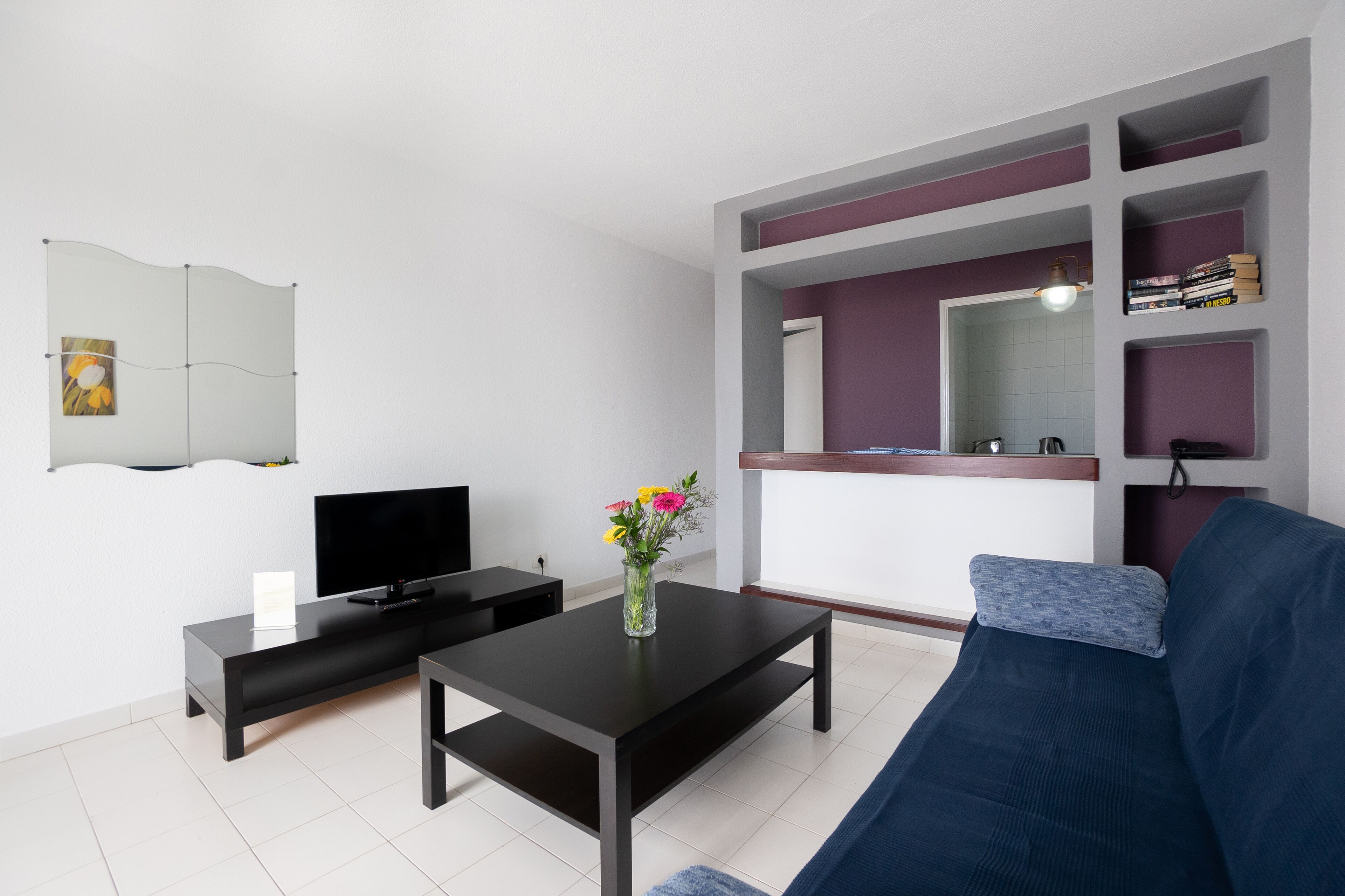 Una sala de estar moderna con un televisor de pantalla plana, una mesa de centro con un jarrón de flores, un sofá oscuro y estanterías empotradas con libros y objetos decorativos.