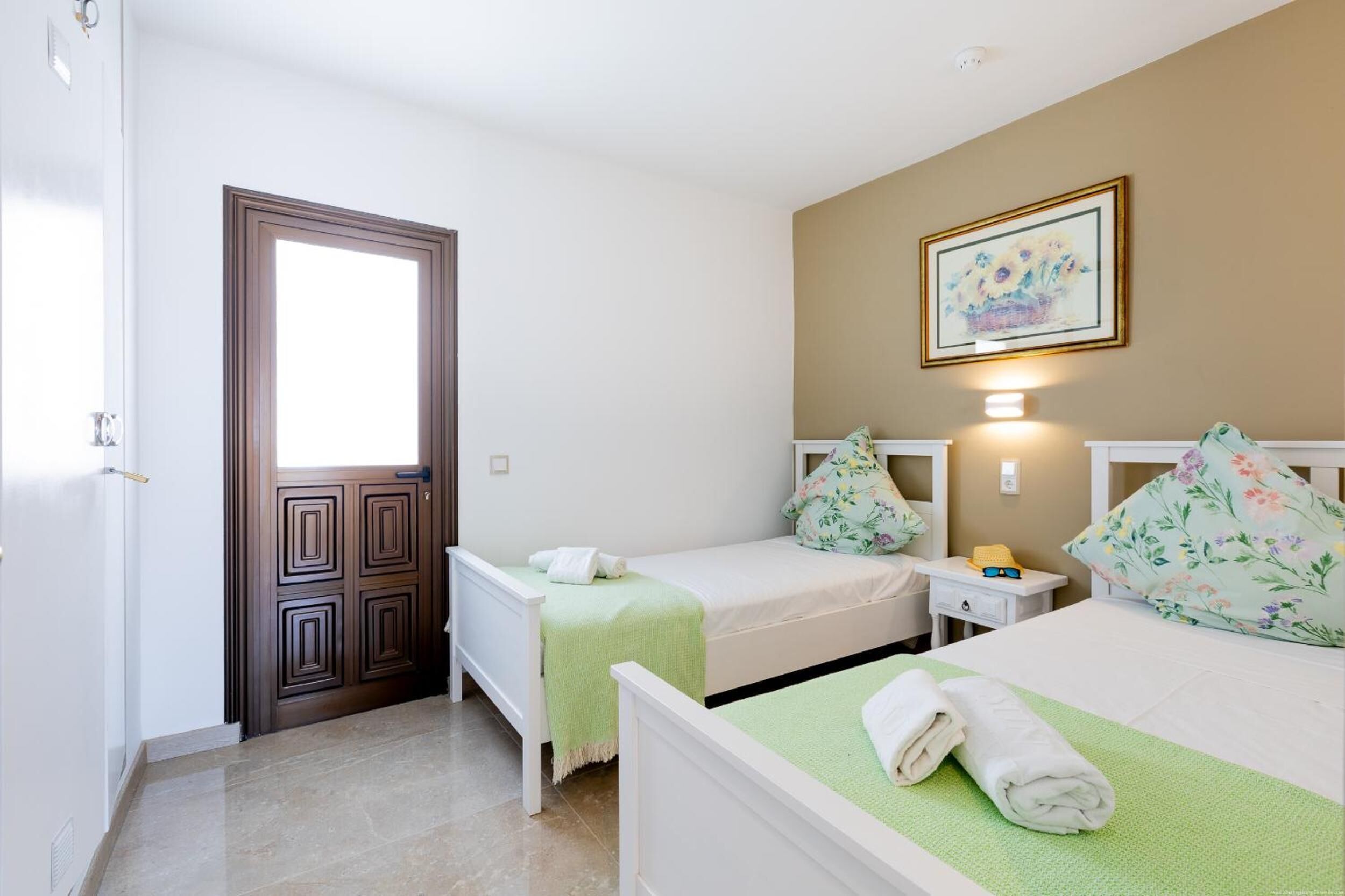 Une chambre d’hôtel avec deux lits, une porte, une lampe fixée au mur et un tableau encadré.