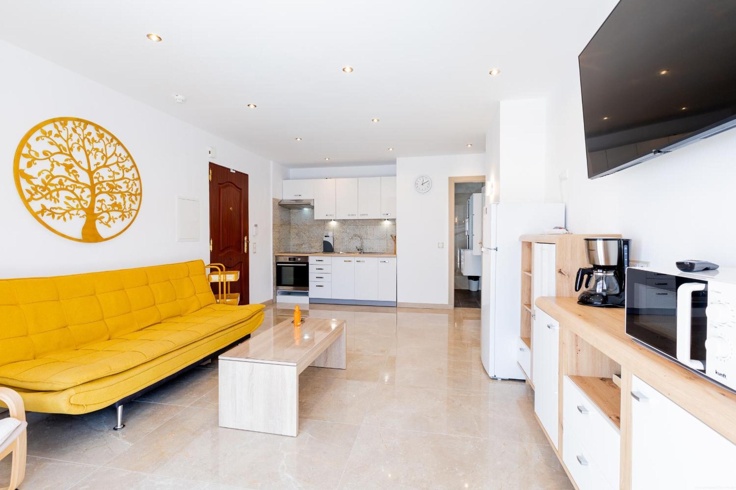 A modern living room with a yellow sofa, a coffee table, a TV mounted on the wall, and a kitchen in the background.