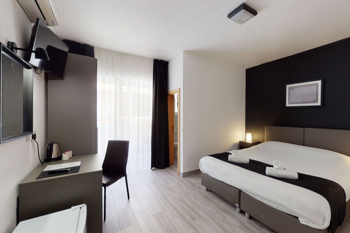 Moderni hotellihuone, jossa on suuri sänky, työpöytä tuolilla, taulutelevisio ja ikkuna, jossa on verhot.