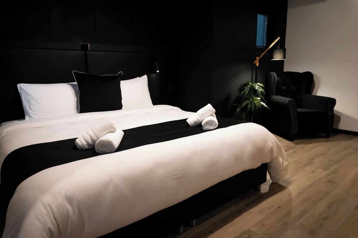 A modern hotel room with a large bed, black and white bedding, a black armchair, a desk lamp, and a potted plant.