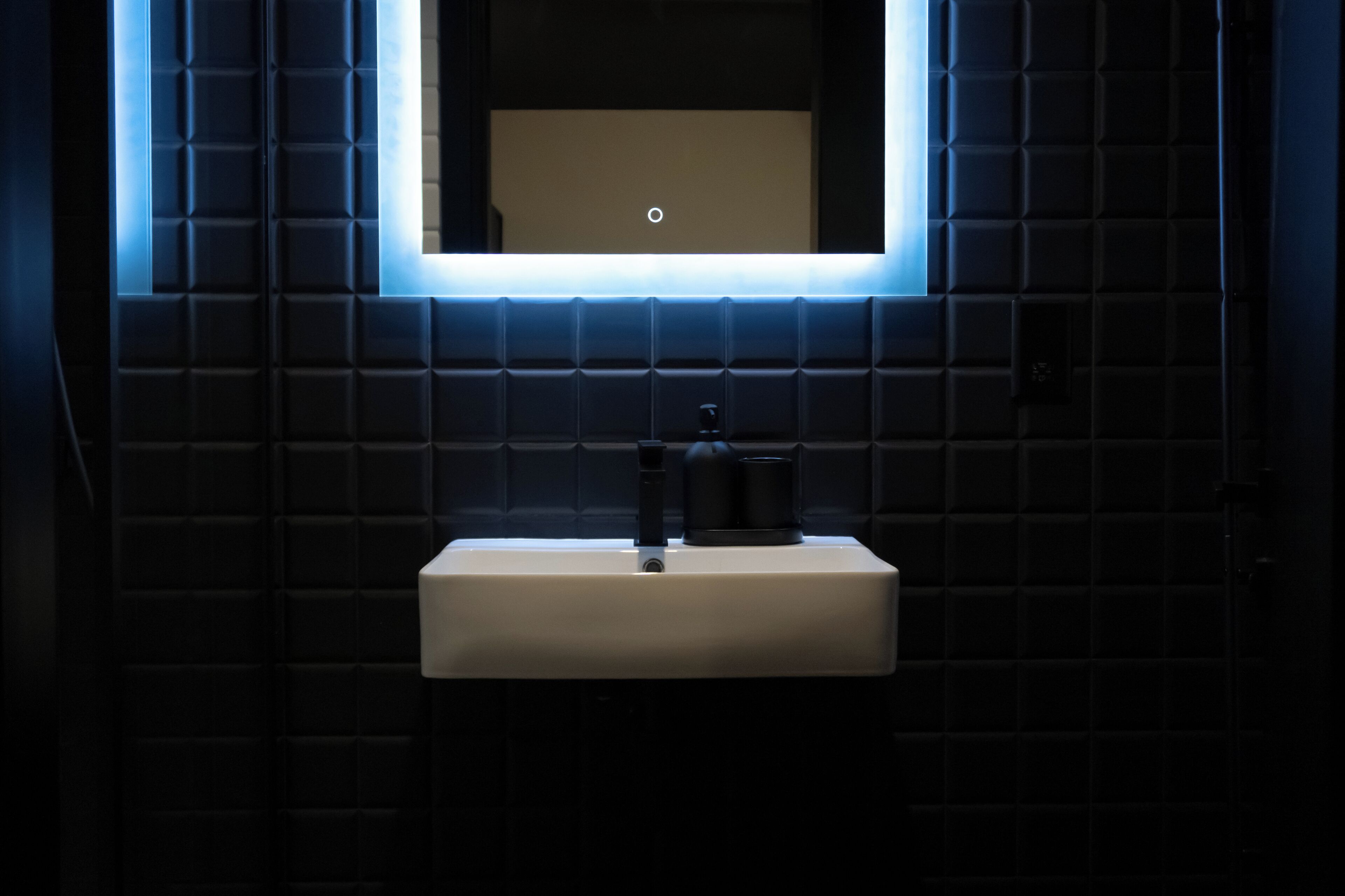 A modern bathroom with a white sink, dark tiled walls, and a rectangular mirror with a lighted frame.
