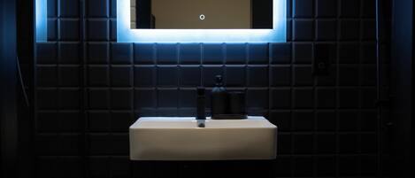 A modern bathroom with a white sink, dark tiled walls, and a rectangular mirror with a lighted frame.