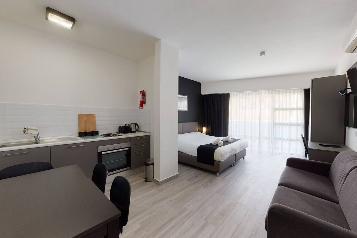 A modern hotel room with a kitchenette, a bed, a sofa, and a TV.