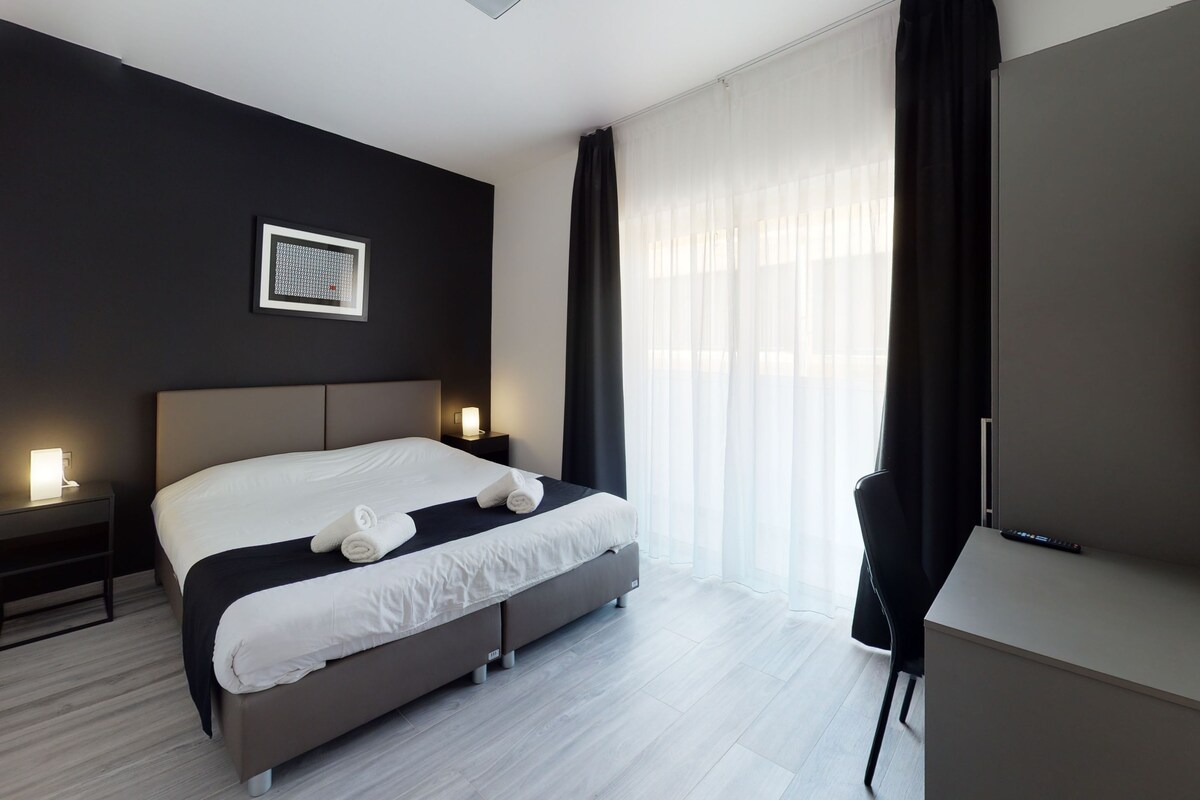 A modern hotel room with a large bed, bedside tables with lamps, a desk, and a chair.