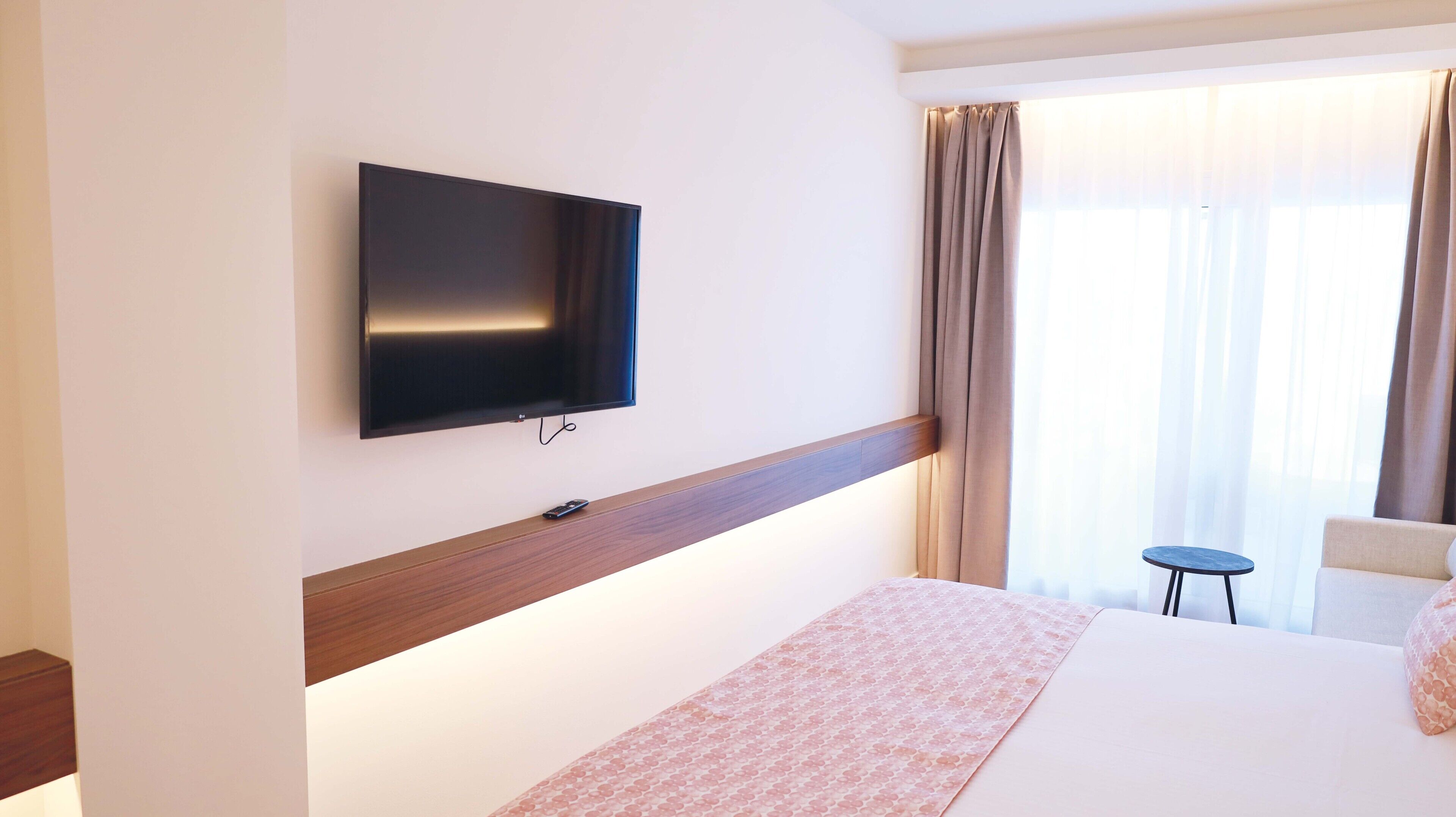 Una habitación de hotel con televisor de pantalla plana montado en la pared, una cama con colcha estampada, una mesita redonda y una silla.