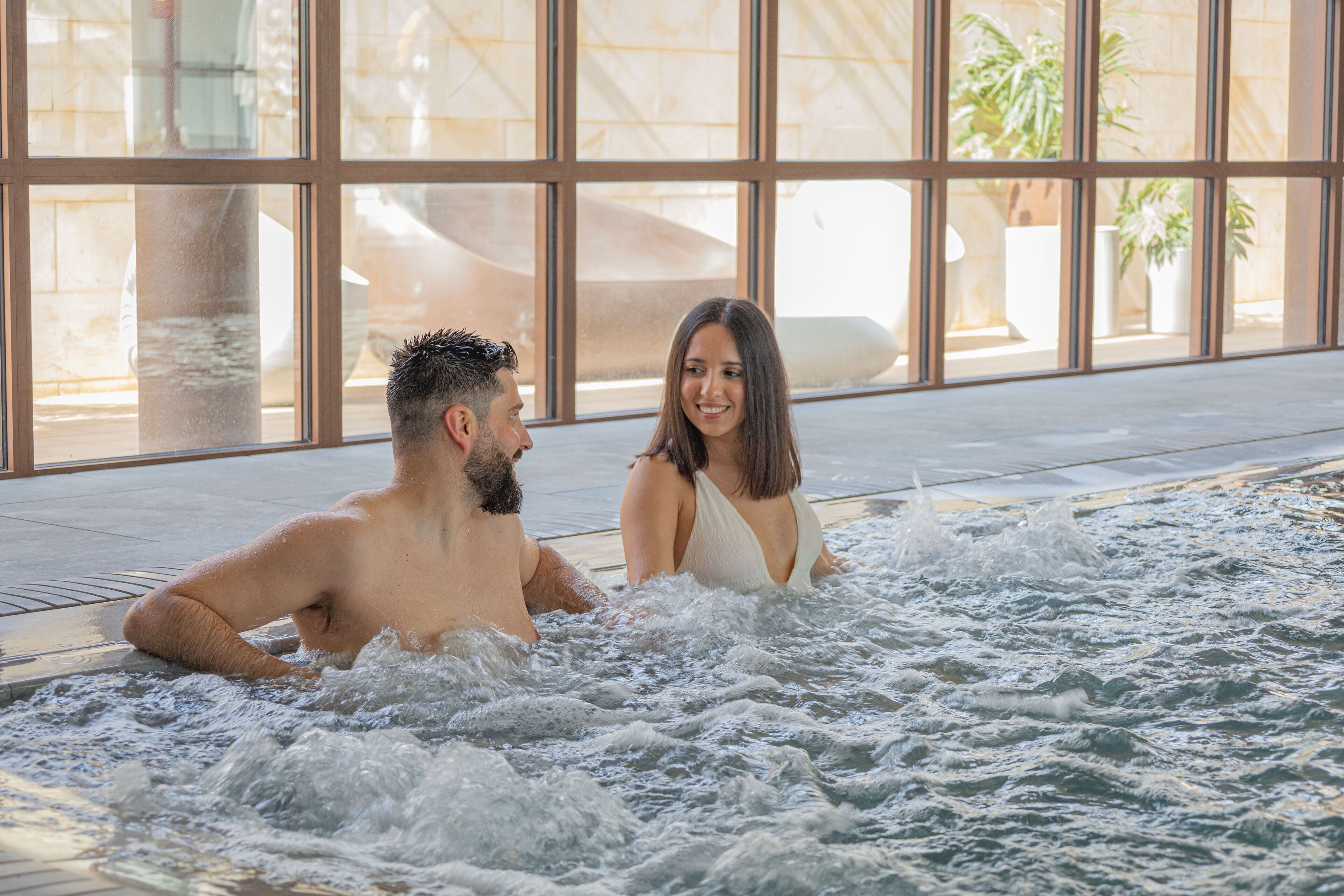 Un couple dans un jacuzzi, avec vue sur un bâtiment moderne et des plantes.