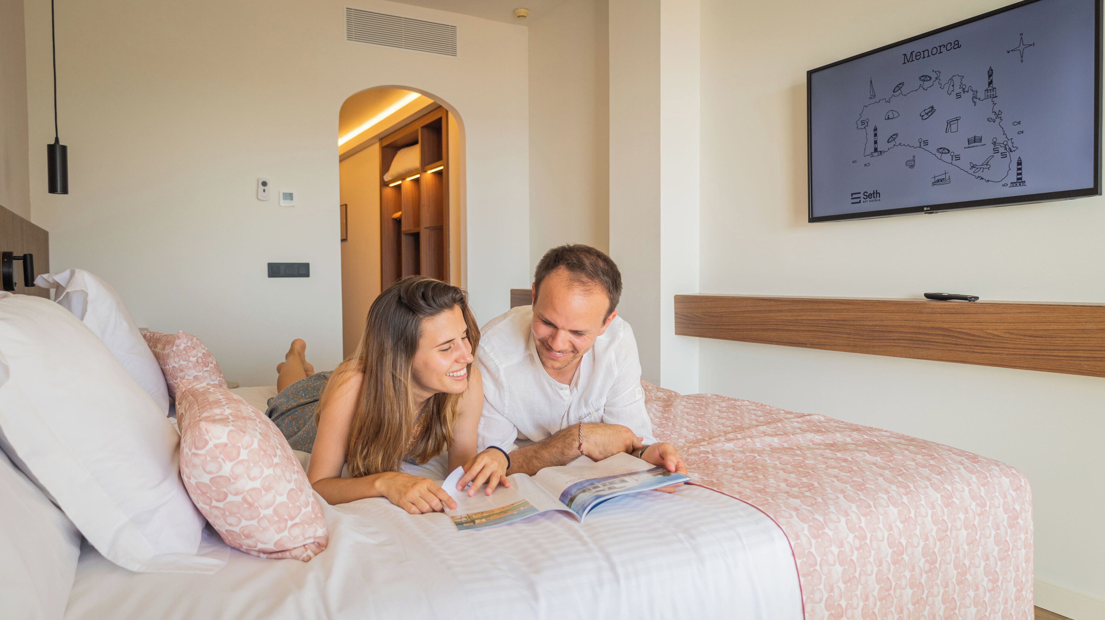 Una pareja leyendo un libro en una cama, con un mapa de Menorca en la pared.