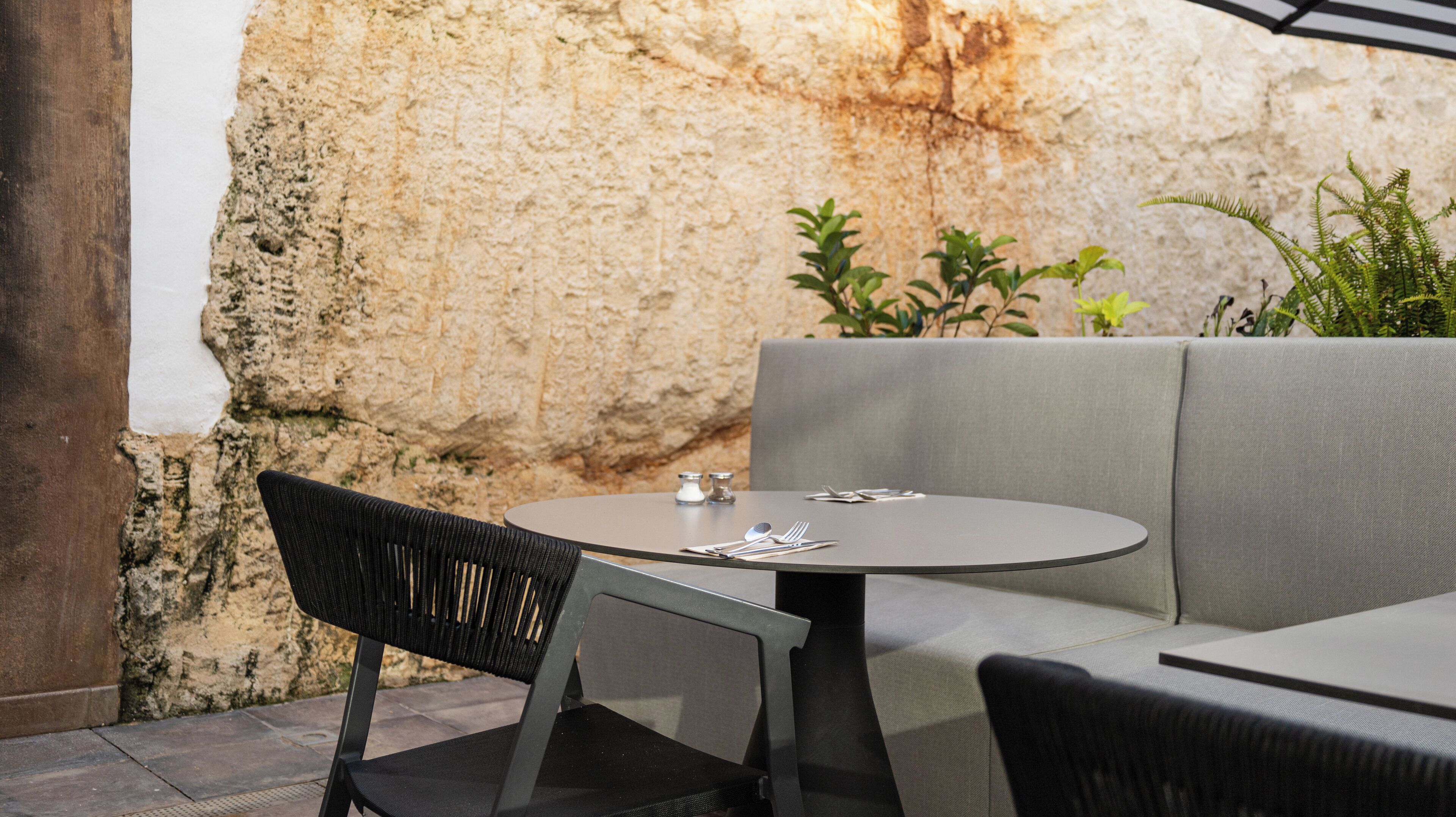 Mesa redonda con un cojín gris, una silla negra y un sofá gris en un entorno exterior con un muro de piedra de fondo.