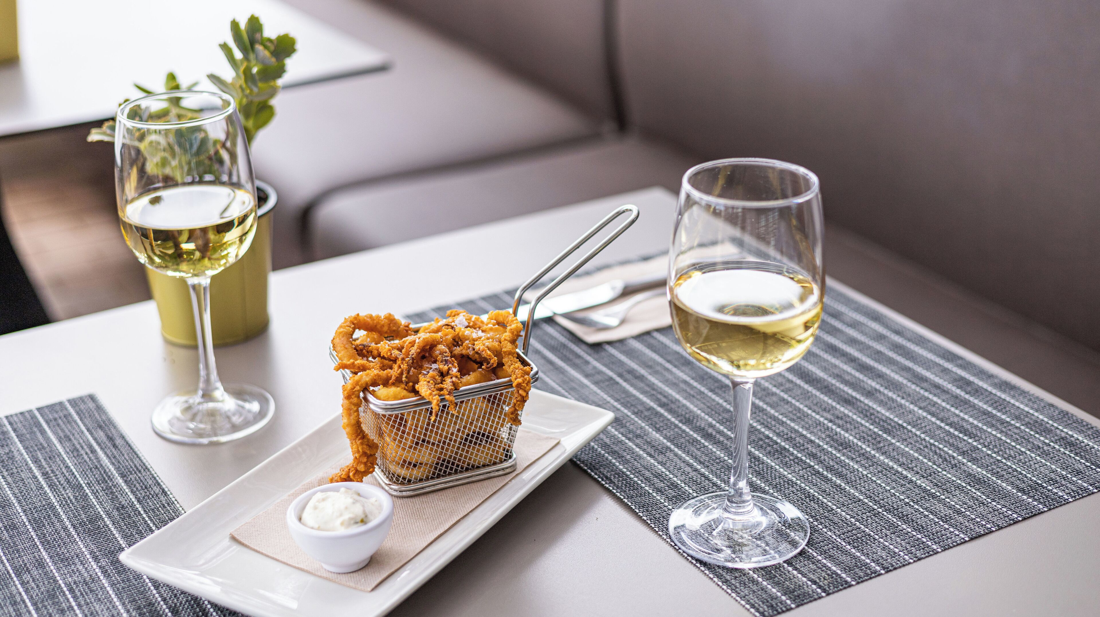 A dining table with a basket of fried food, a small bowl of sauce, and two glasses of white wine.
