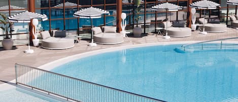 Zona de piscina con tumbonas y sombrillas, rodeada de paredes de cristal.