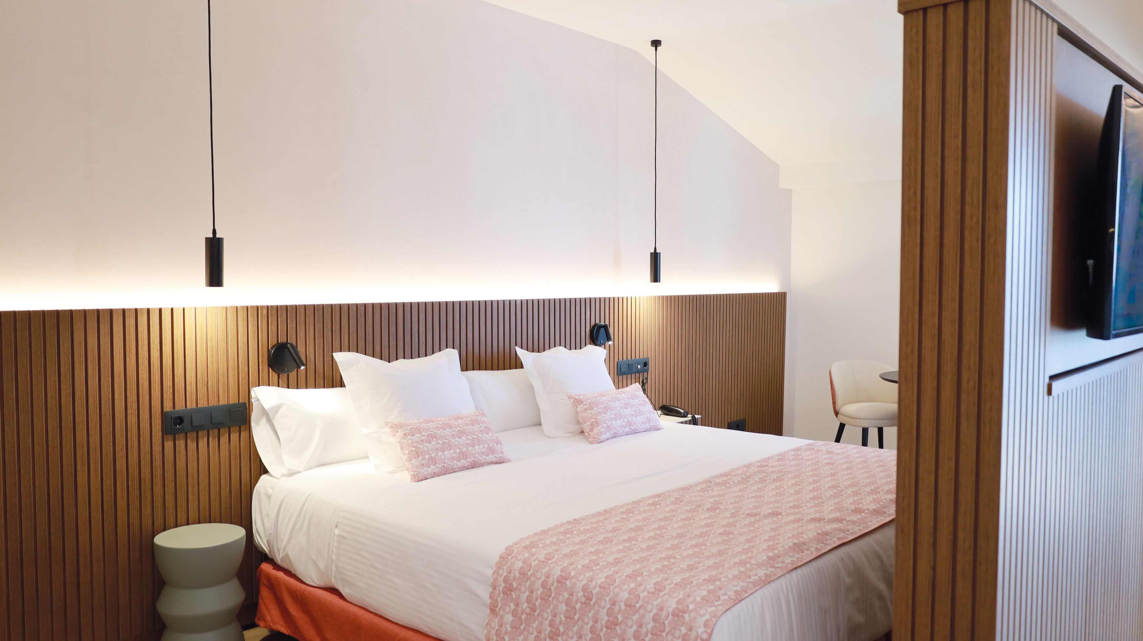 Una habitación de hotel moderna con una cama grande, paneles de madera en las paredes, un televisor de pantalla plana montado y una pequeña mesa redonda con una silla.
