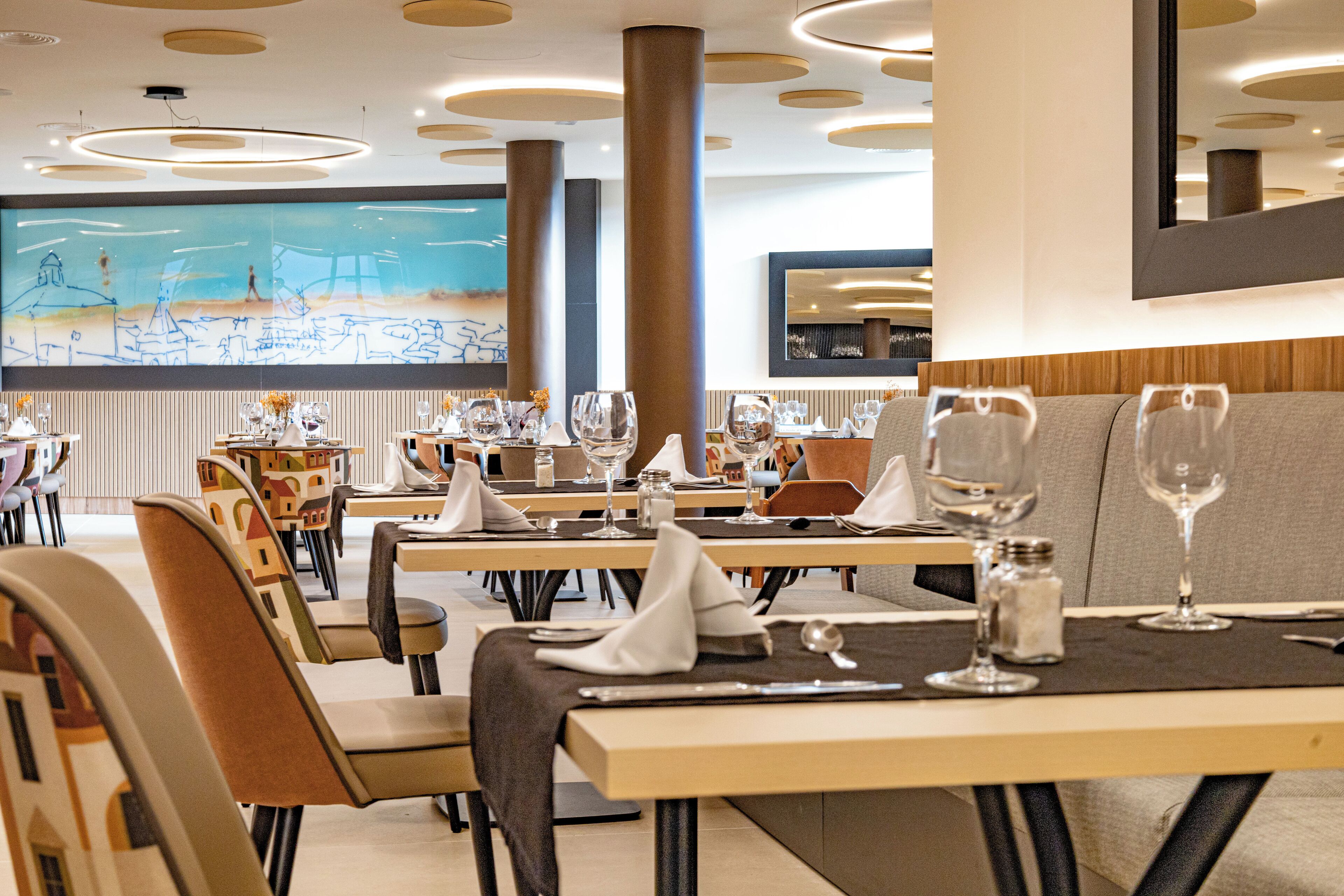 A modern dining area with tables set for two, featuring contemporary chairs and a large abstract mural on the wall.