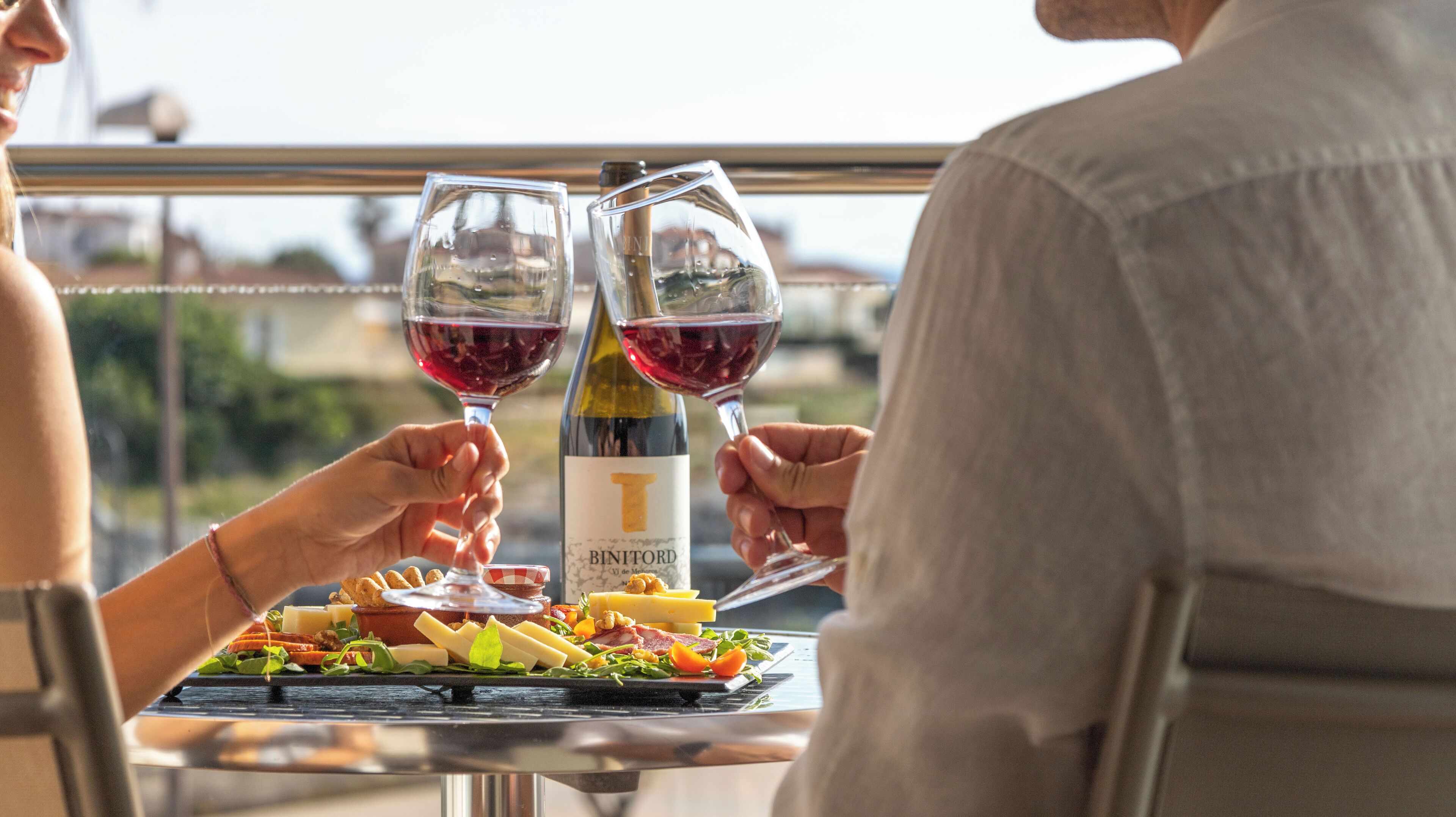 A table with a bottle of wine and two glasses of red wine, a plate of food, and a scenic view through a window.