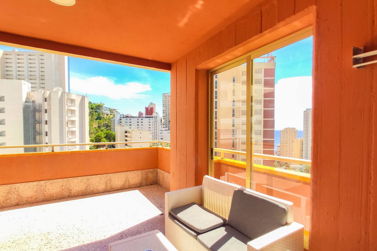 A balcony with a white table and two wicker chairs, overlooking a cityscape with tall buildings.