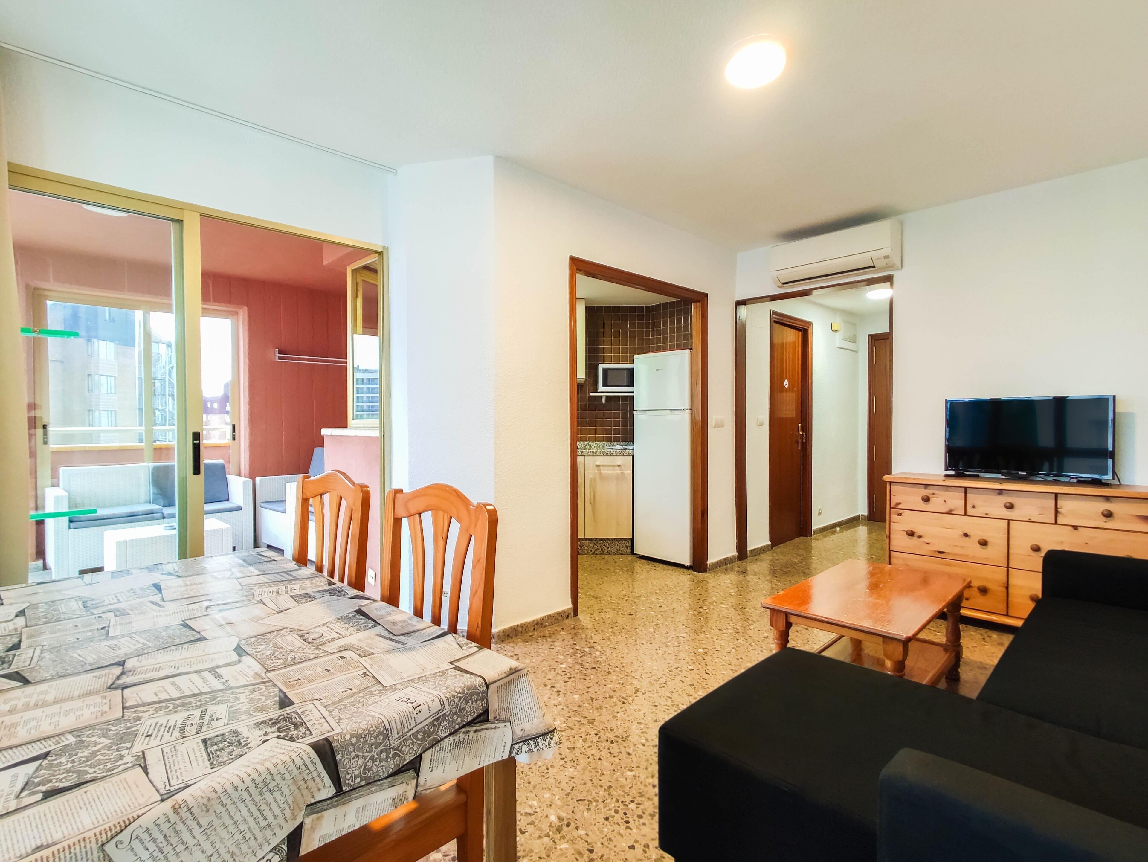 A room with a dining table, chairs, a sofa, a TV, and a kitchen area with a refrigerator and microwave.