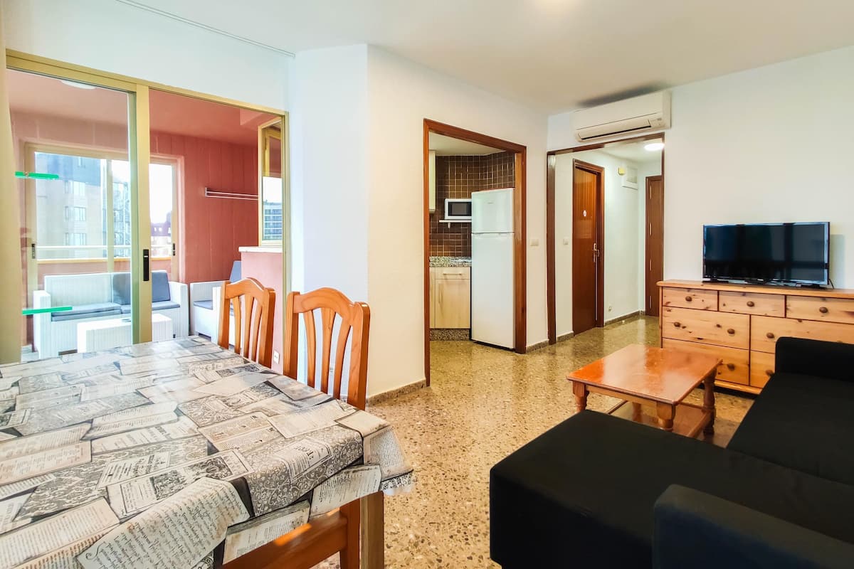 A room with a dining table, chairs, a sofa, a TV, and a kitchen area with a refrigerator and microwave.