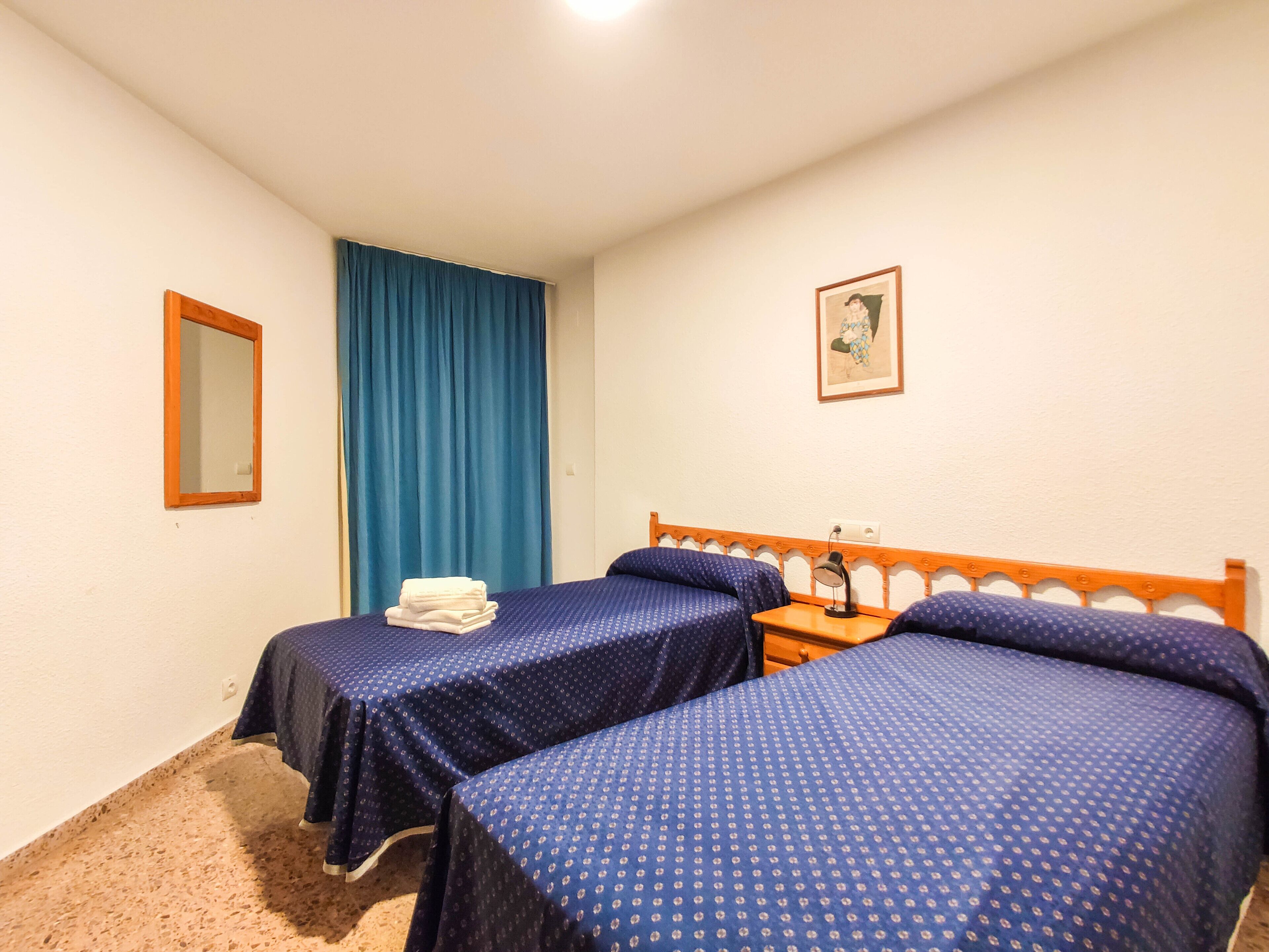 A hotel room with two beds, a mirror, a blue curtain, a framed picture, and a small table with a lamp.