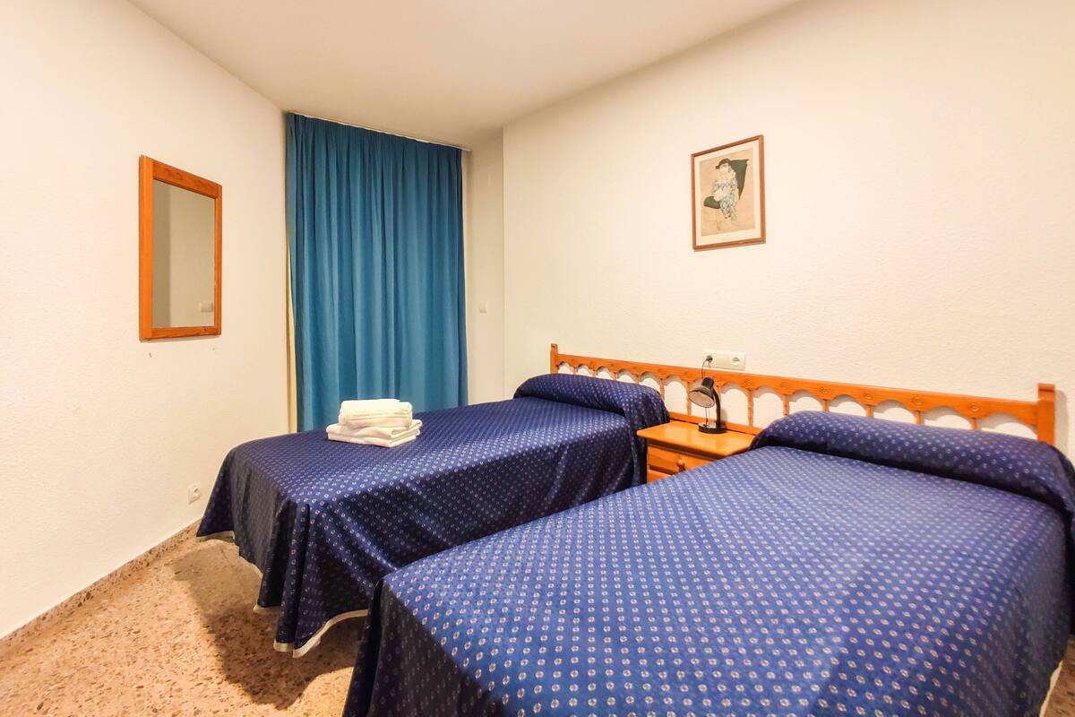 A hotel room with two beds, a mirror, a blue curtain, a framed picture, and a small table with a lamp.