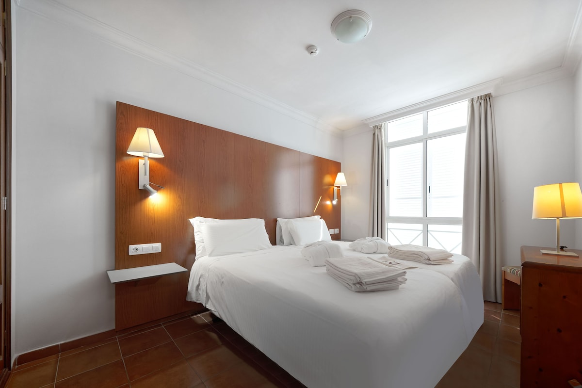 A hotel room with a large bed, two wall-mounted lamps, a wooden headboard, a window with curtains, and a mirror.