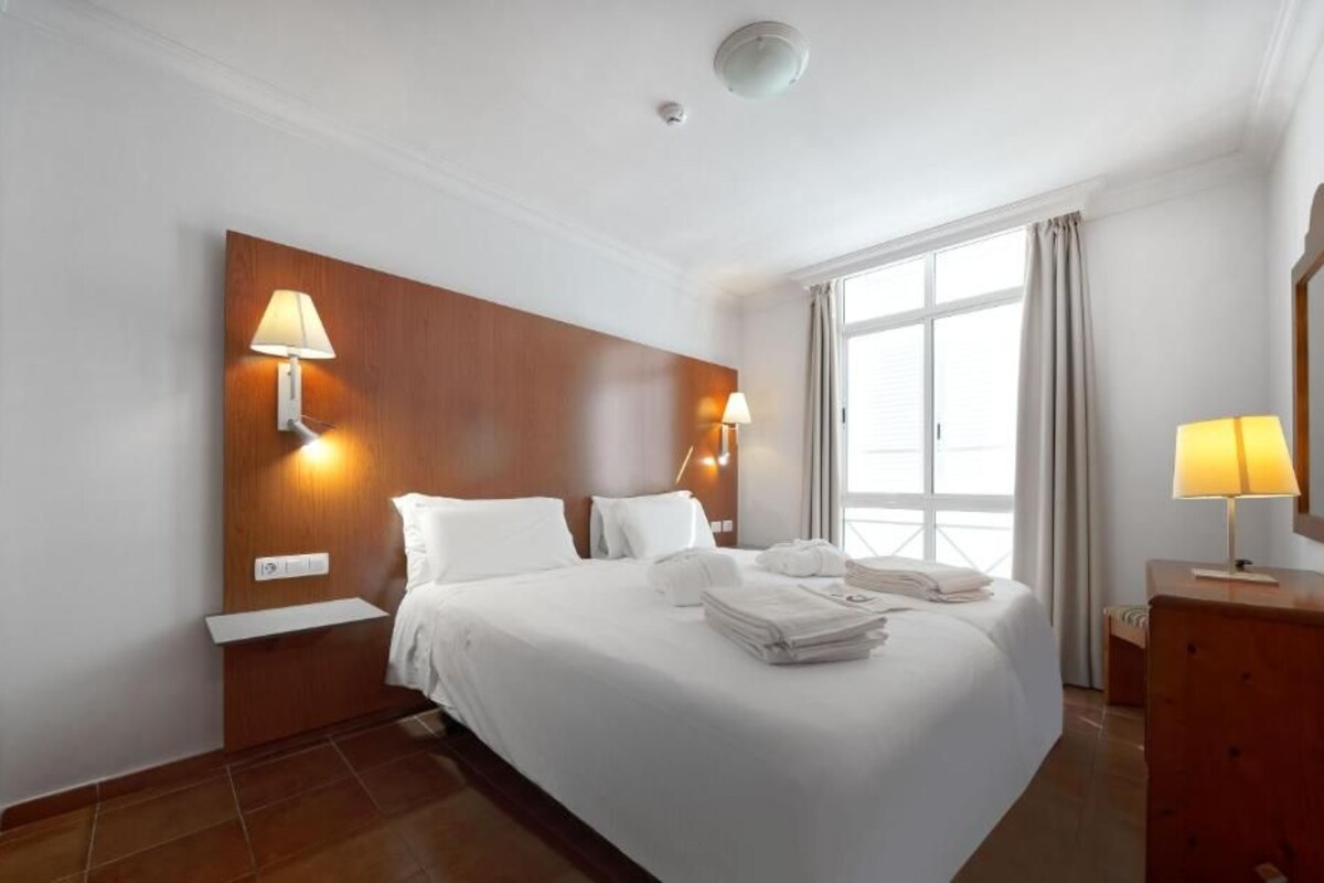 A hotel room with a large bed, two bedside lamps, a wooden headboard, a nightstand, a dresser, and a window with curtains.