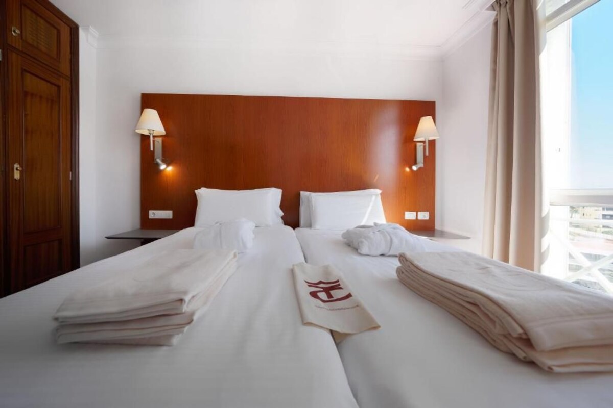 A hotel room with a wooden headboard, two bedside lamps, neatly folded towels, and a view of the city through the window.
