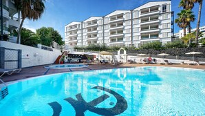 A hotel with a swimming pool featuring a large logo, a covered seating area, and a view of the building's balconies. - Apartamentos Dorotea (San Bartolomé de Tirajana)