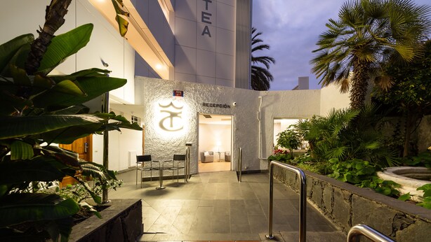 A hotel entrance with a reception desk, a sign with a symbol, and a stone wall with plants. - Apartamentos Dorotea (San Bartolomé de Tirajana)