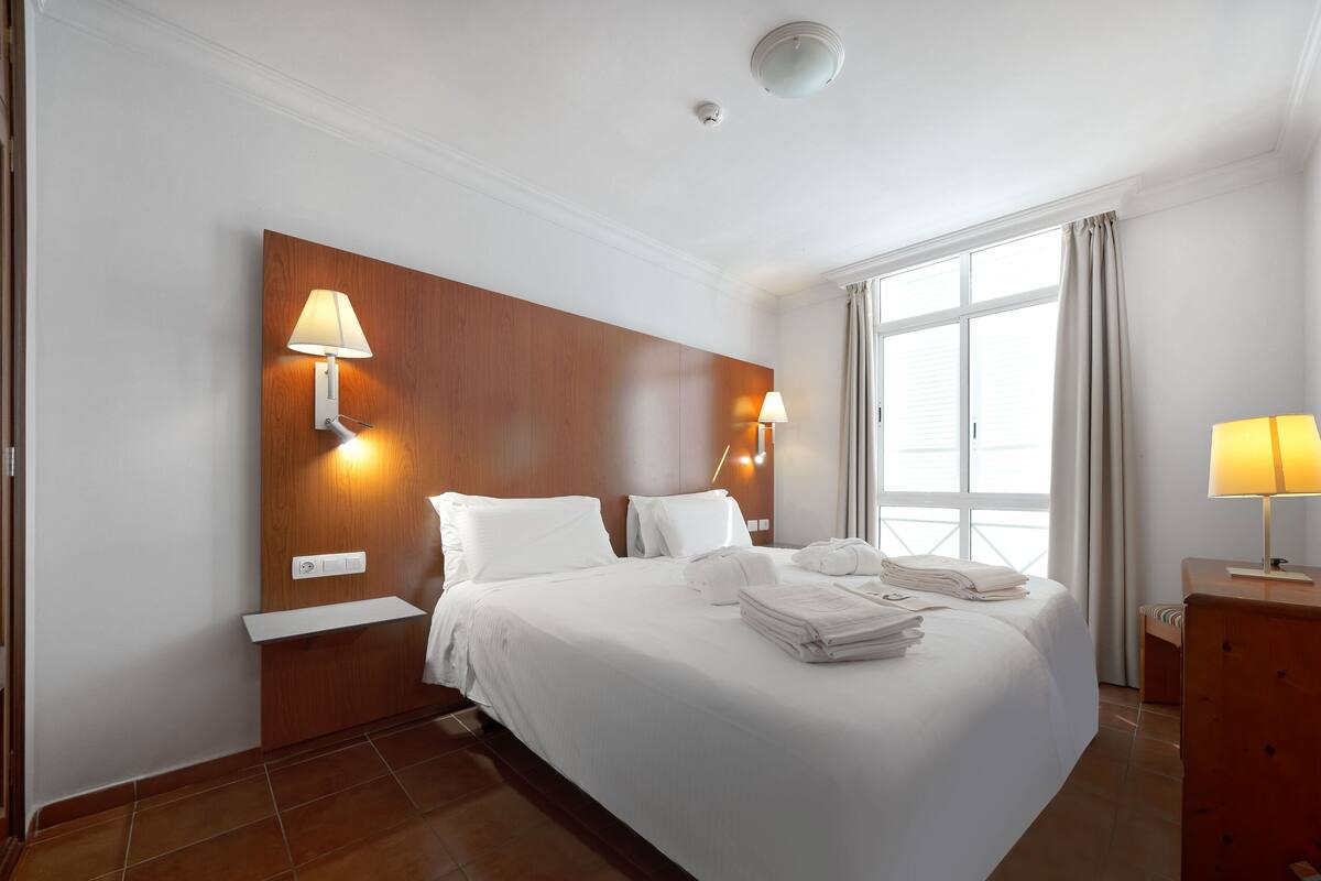 A hotel room with a large bed, two wall-mounted lamps, a wooden headboard, a window with curtains, and a mirror.