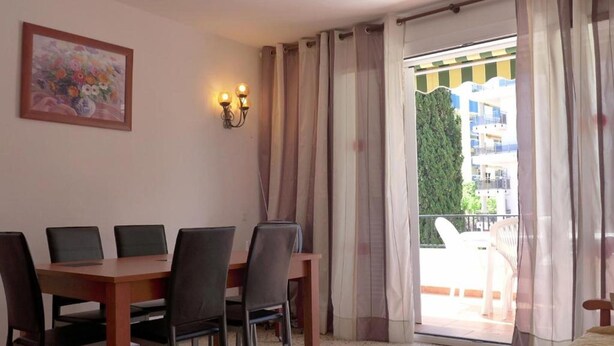 Ein Zimmer mit Esstisch, Stühlen, einem Bett, einem Gemälde und einem Balkon mit Vorhängen.