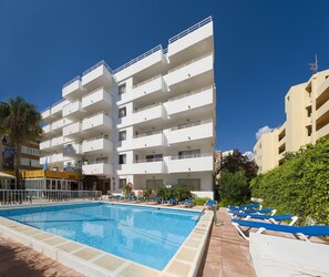 A poolside area with a rectangular pool, white multi-story buildings, and blue lounge chairs. - Green Line Bon Sol Apartamentos (Ibiza Town)