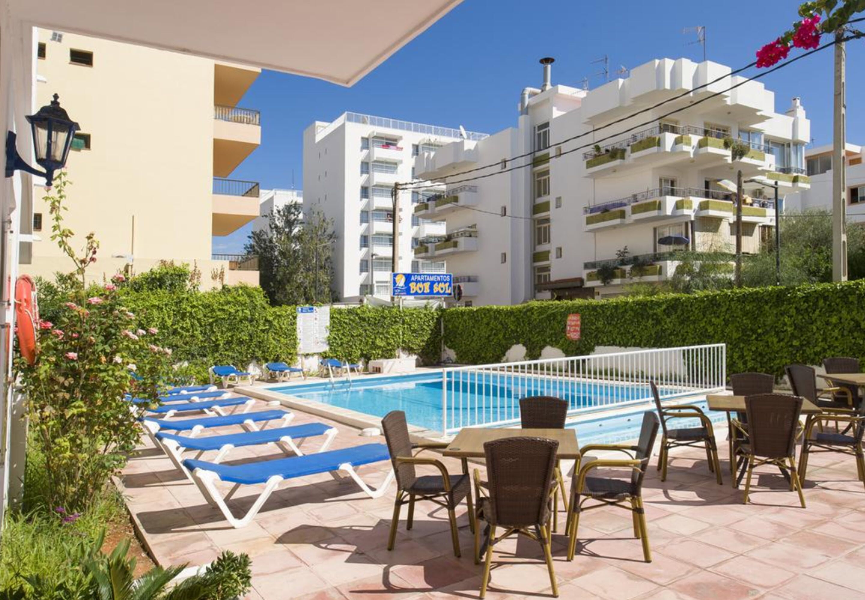 Zona de piscina con sillas reclinables y mesas, rodeada de follaje y un edificio con balcones.