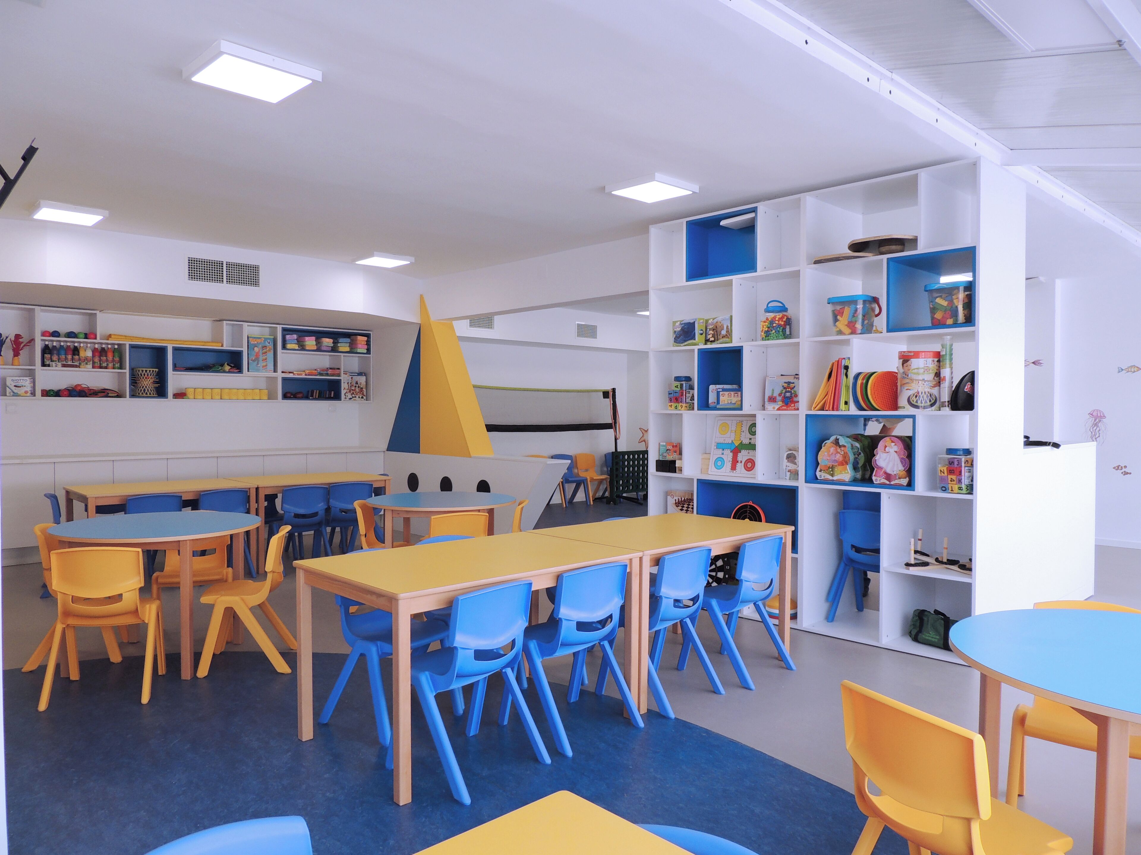 Un cuarto de juegos infantil con mesas y sillas, estanterías llenas de juguetes y material educativo.