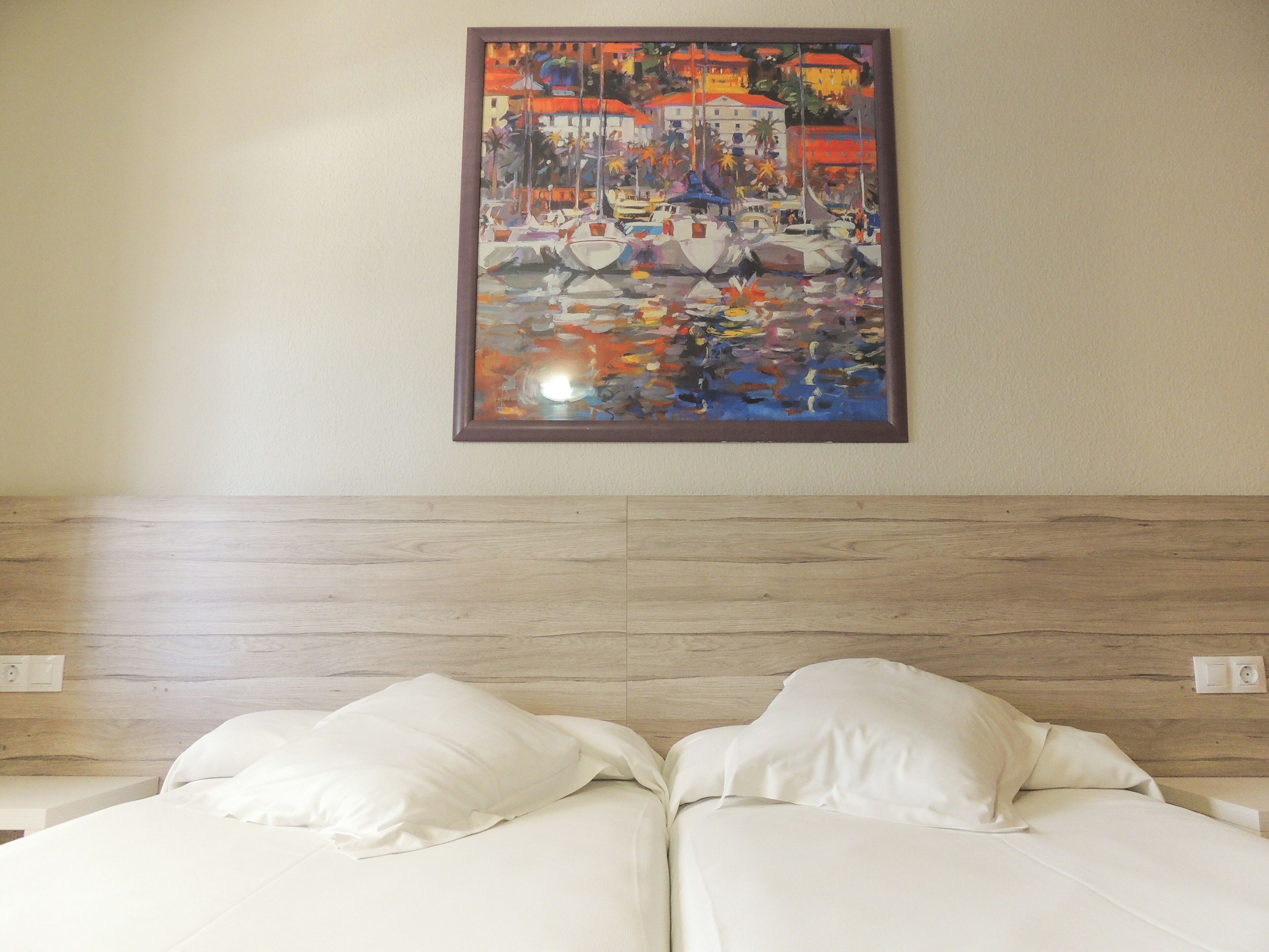 Une chambre d’hôtel avec un lit, deux oreillers, une tête de lit en bois et un tableau coloré accroché au mur.