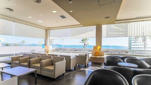 A modern lounge area with seating and tables, large windows with a view of the sea, and a balcony with a railing.
