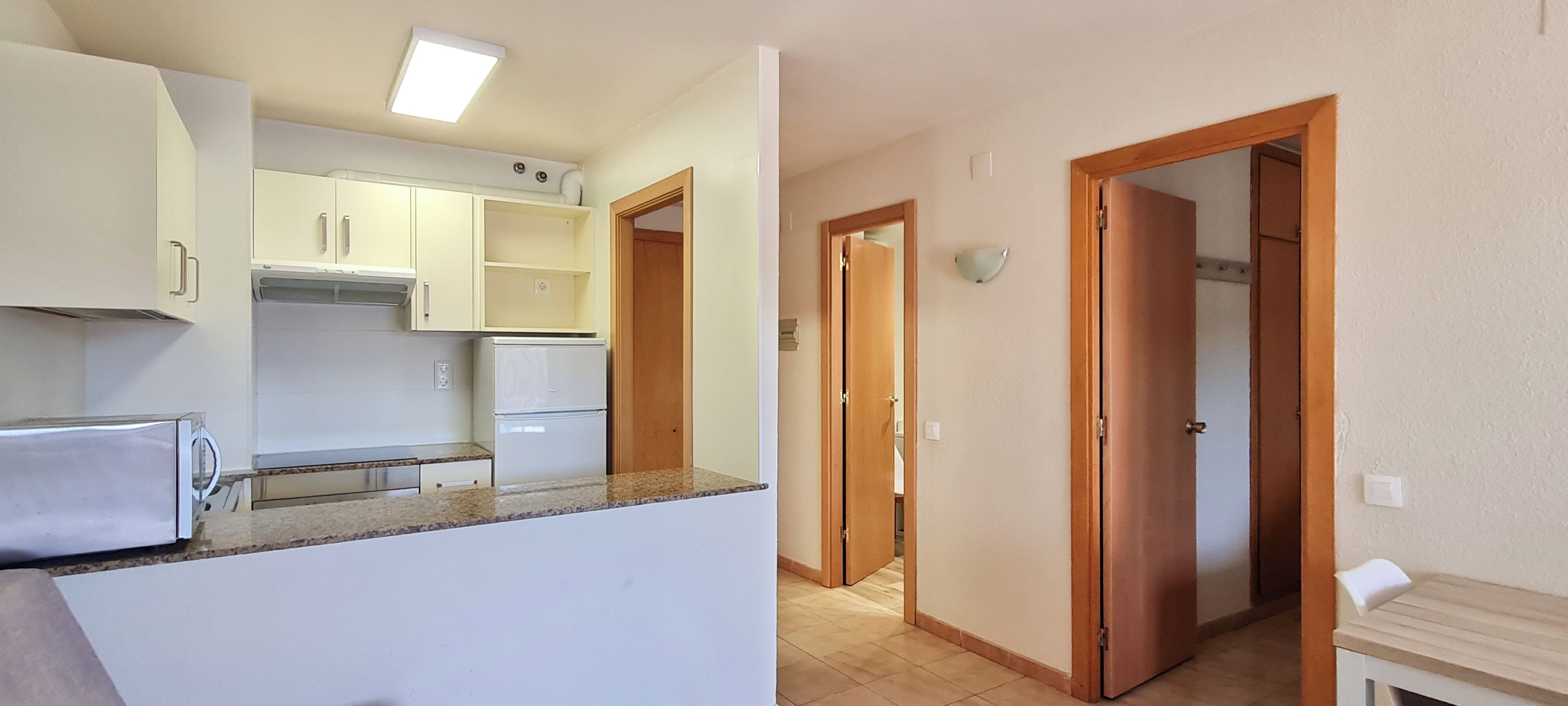 Una cucina con mobili bianchi, un piano di lavoro e un frigorifero. Si apre una porta in legno verso un'altra stanza.