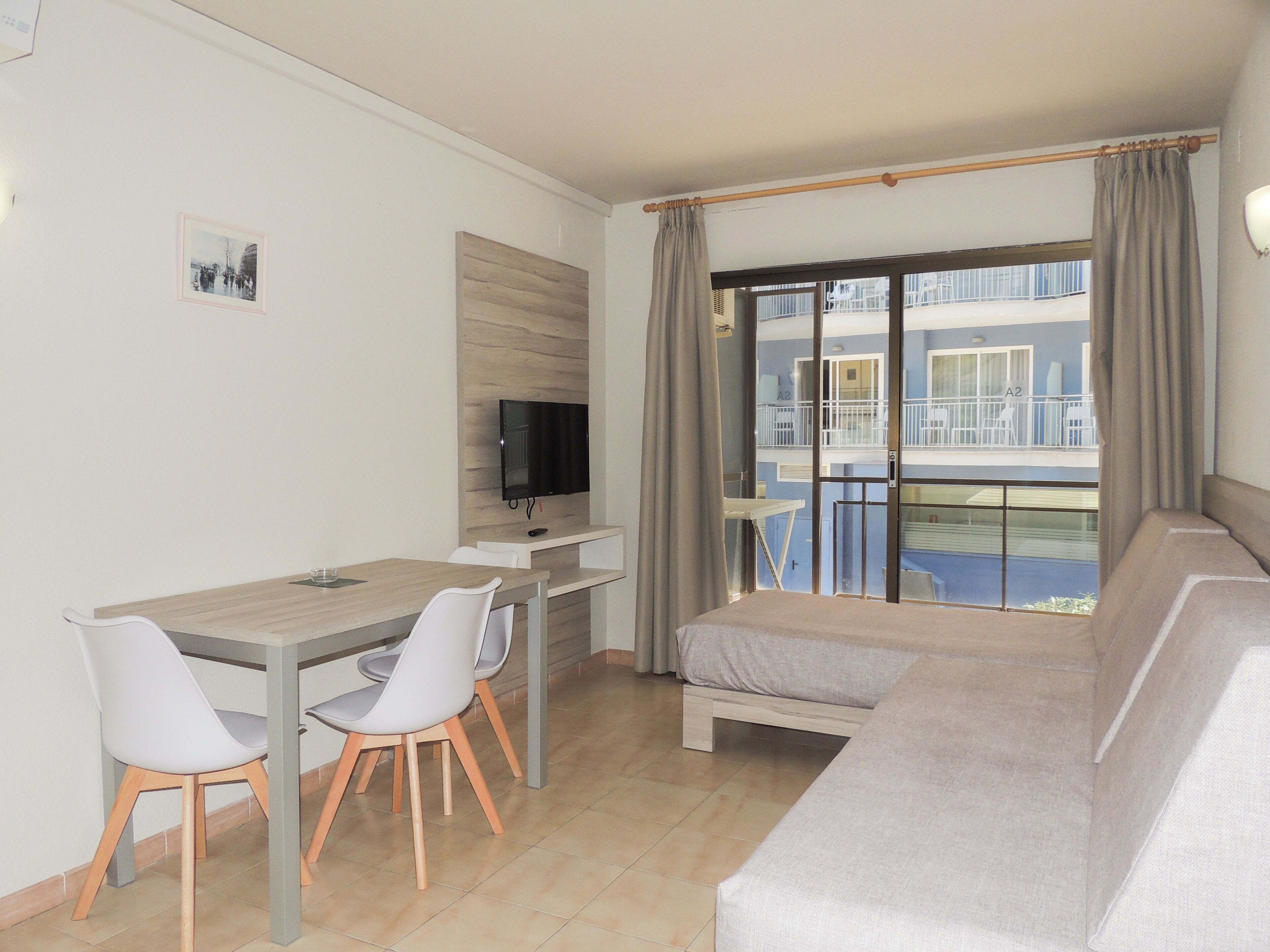 A modern living room with a sofa, a dining table with chairs, a television mounted on the wall, and a balcony with a view of the outside.