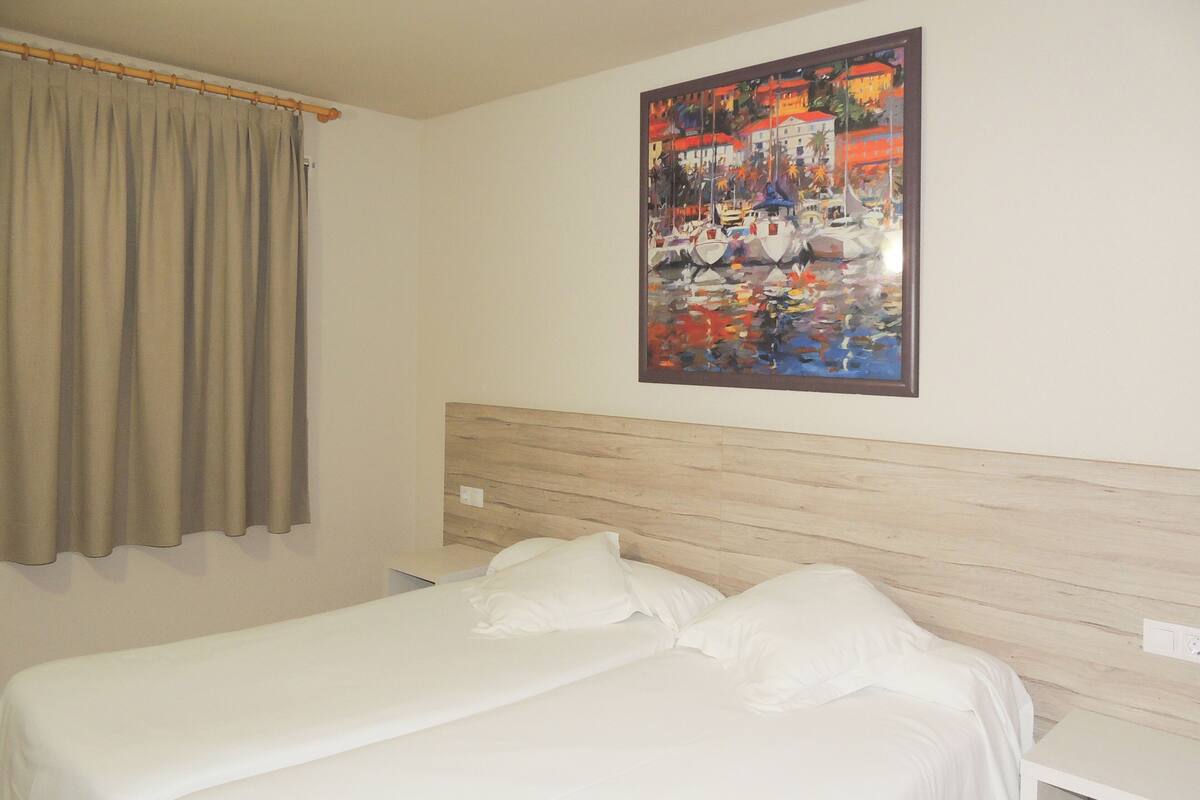 A hotel room with a bed, a nightstand, a painting on the wall, and a window with curtains.