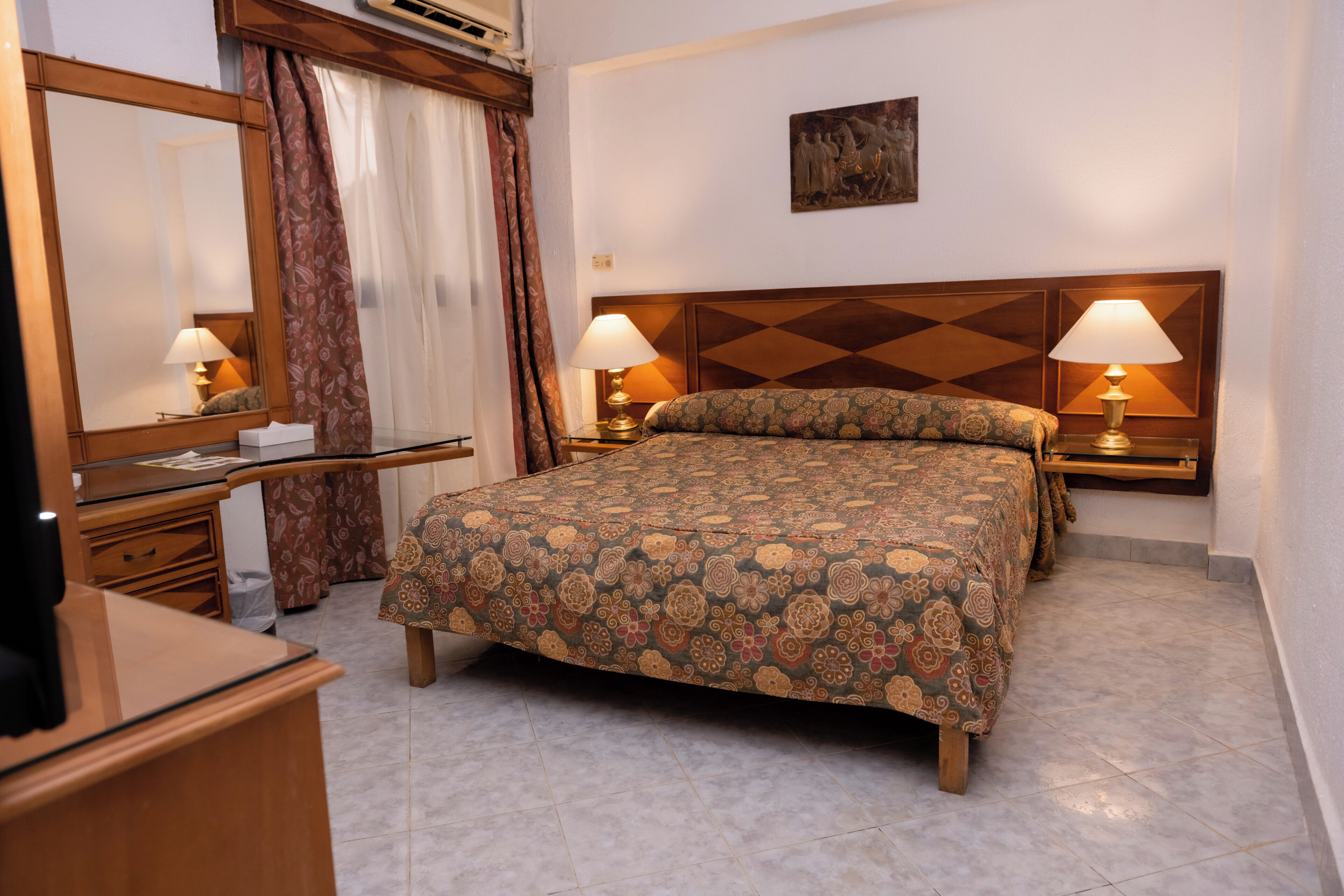 A hotel room with a bed, two bedside lamps, a dresser, a mirror, and a painting on the wall.