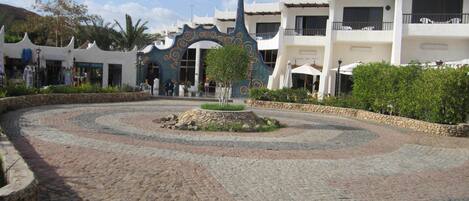 A courtyard with cobblestone paving, a central decorative fountain, and buildings with balconies and outdoor seating.