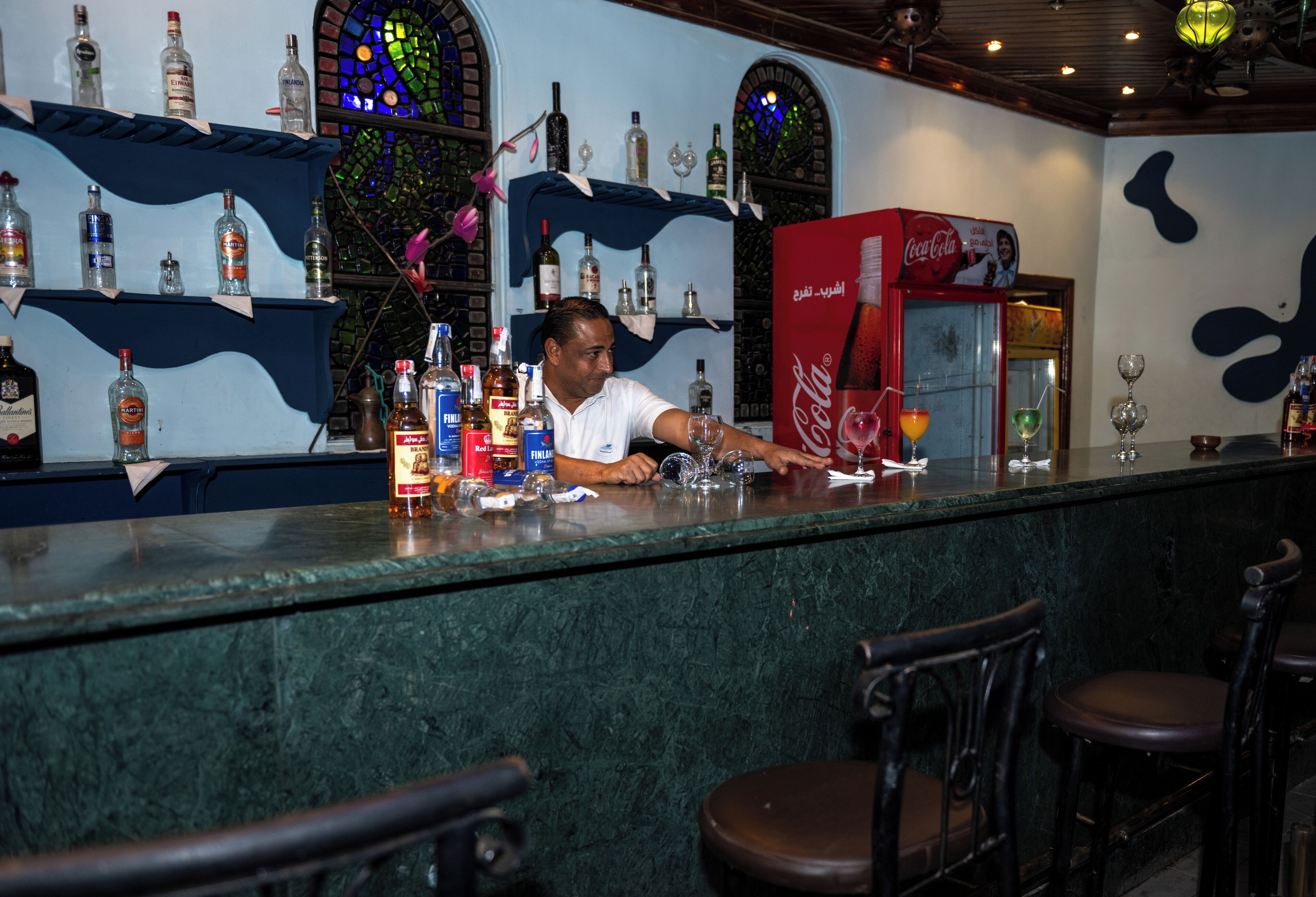 A bar area with a bartender, shelves with liquor bottles, a Coca-Cola refrigerator, and bar stools.