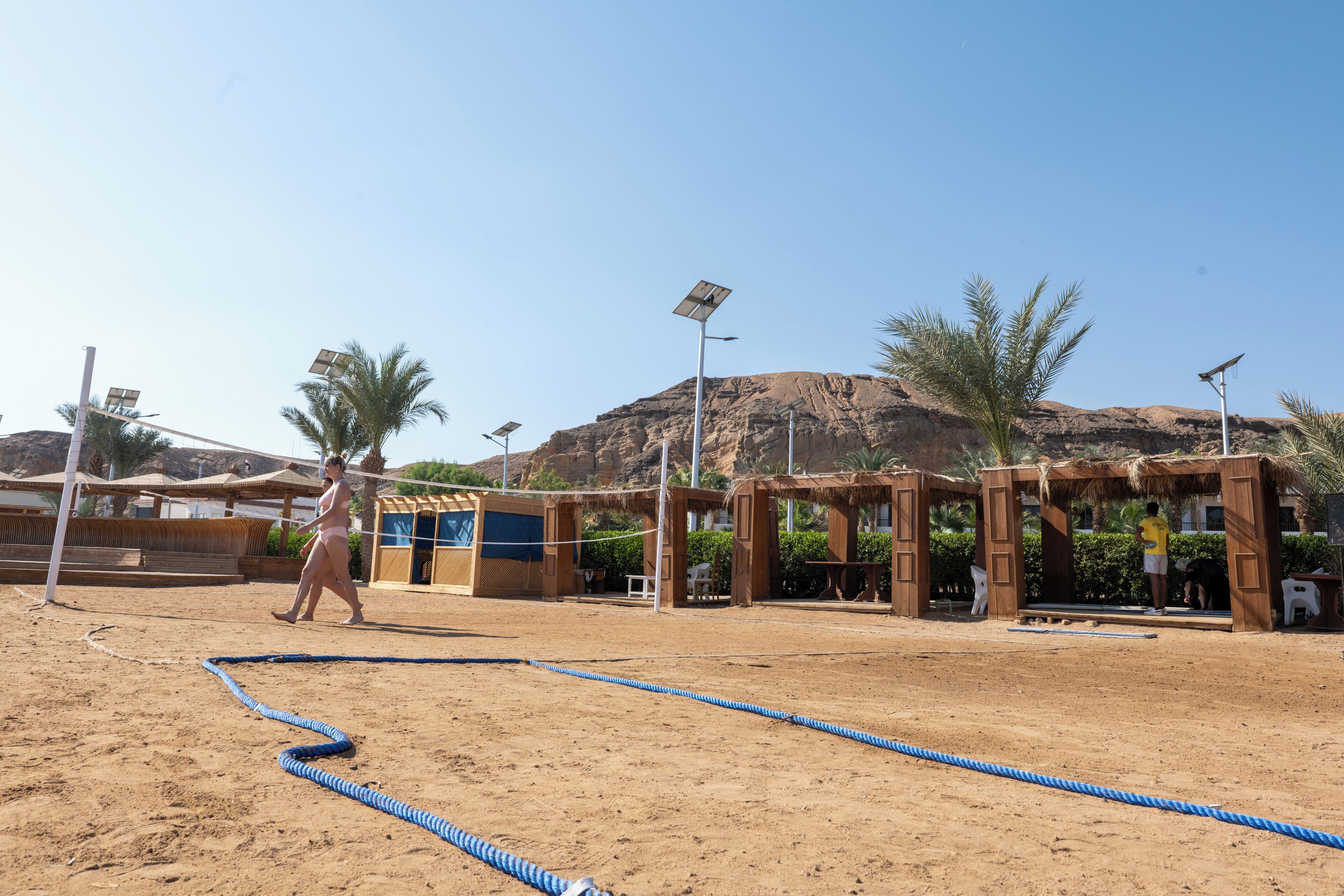 A sandy outdoor area with a volleyball net, palm trees, and a mountainous backdrop.