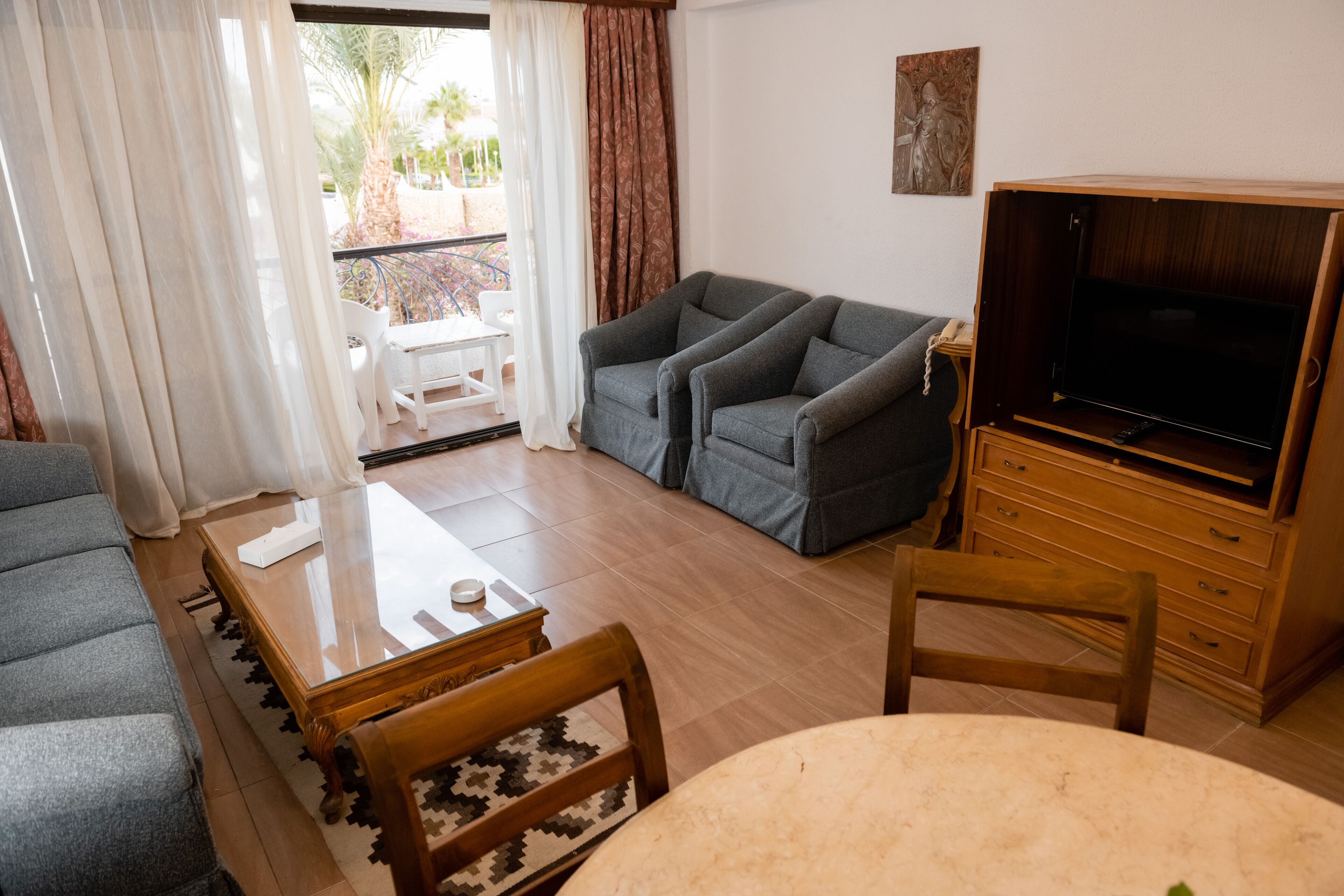 Un salon comprenant un canapé, un téléviseur, une table à manger avec des chaises et un balcon offrant une vue sur les palmiers.