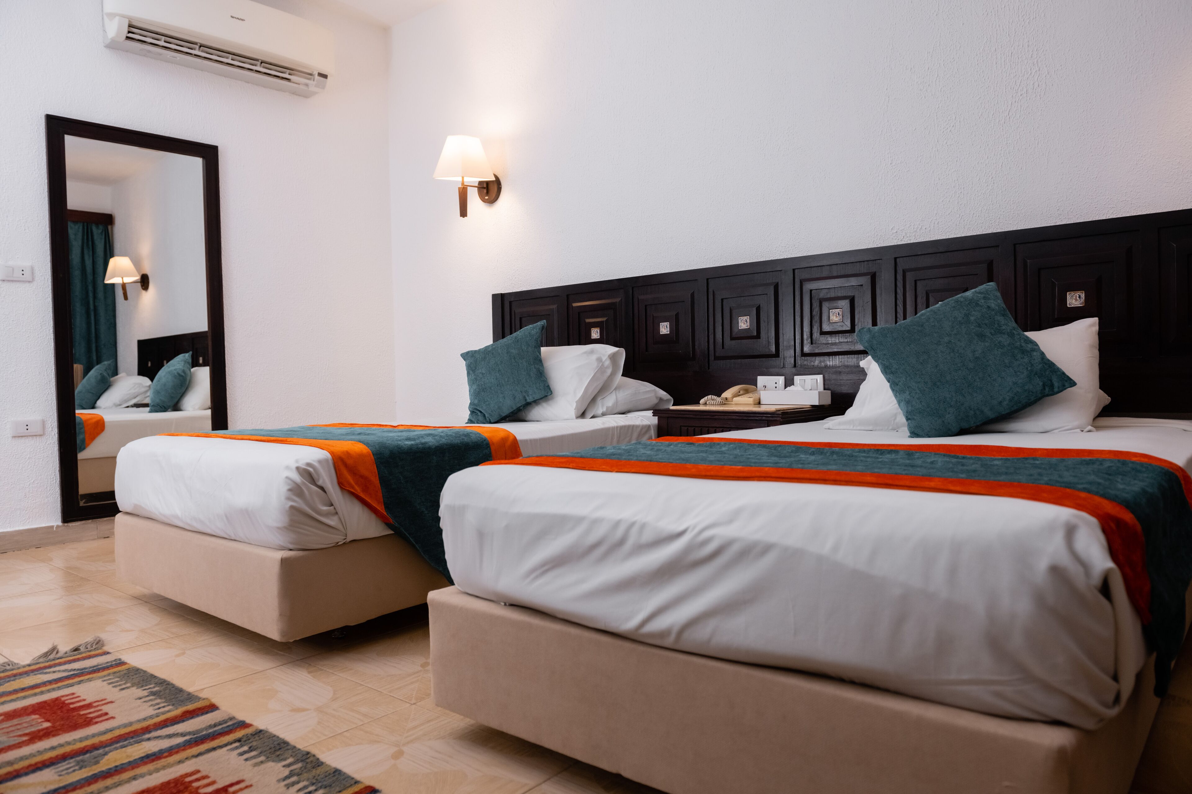 Une chambre d’hôtel avec deux lits, un grand miroir, une lampe fixée au mur et un sol carrelé.