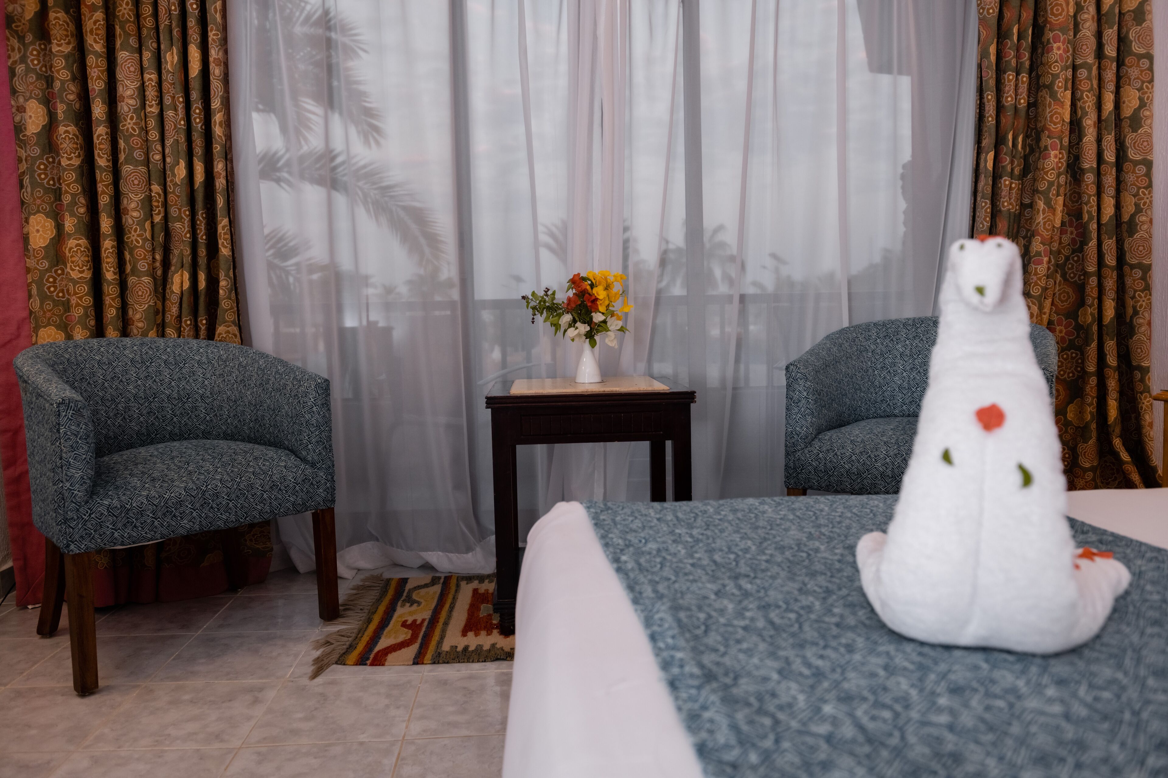 A hotel room with a bed, two patterned armchairs, a small table with a vase of flowers, and curtains with a floral pattern.