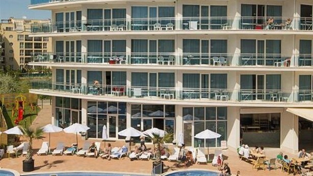 Un hotel de varios pisos con zona de piscina, mobiliario exterior y un cielo azul despejado.