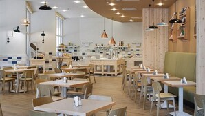 Un restaurante con mesas y sillas de madera, iluminación colgante y una zona de cocina al fondo.